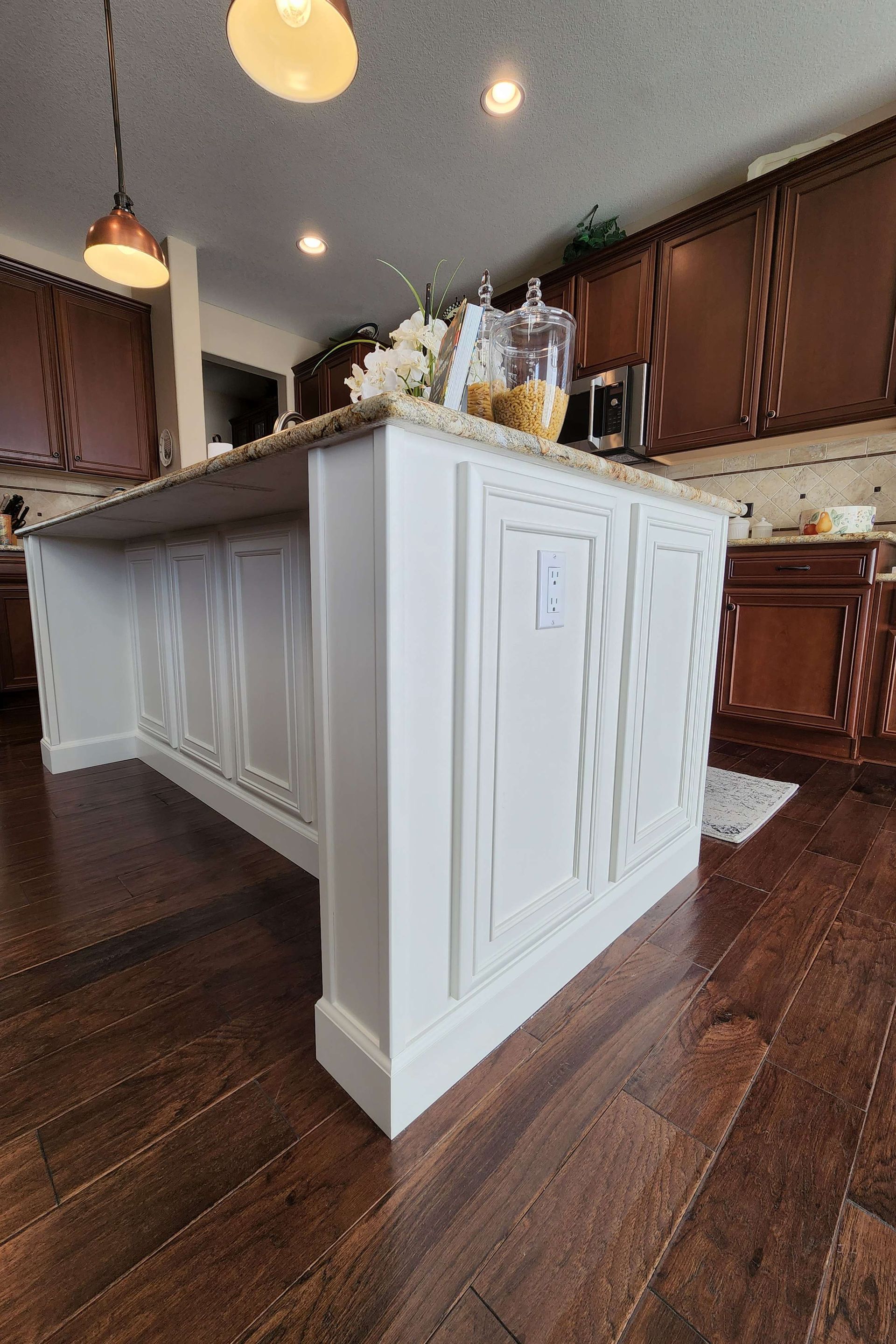 White kitchen island with decorative panels, dark wood floor, brown cabinets in the background.