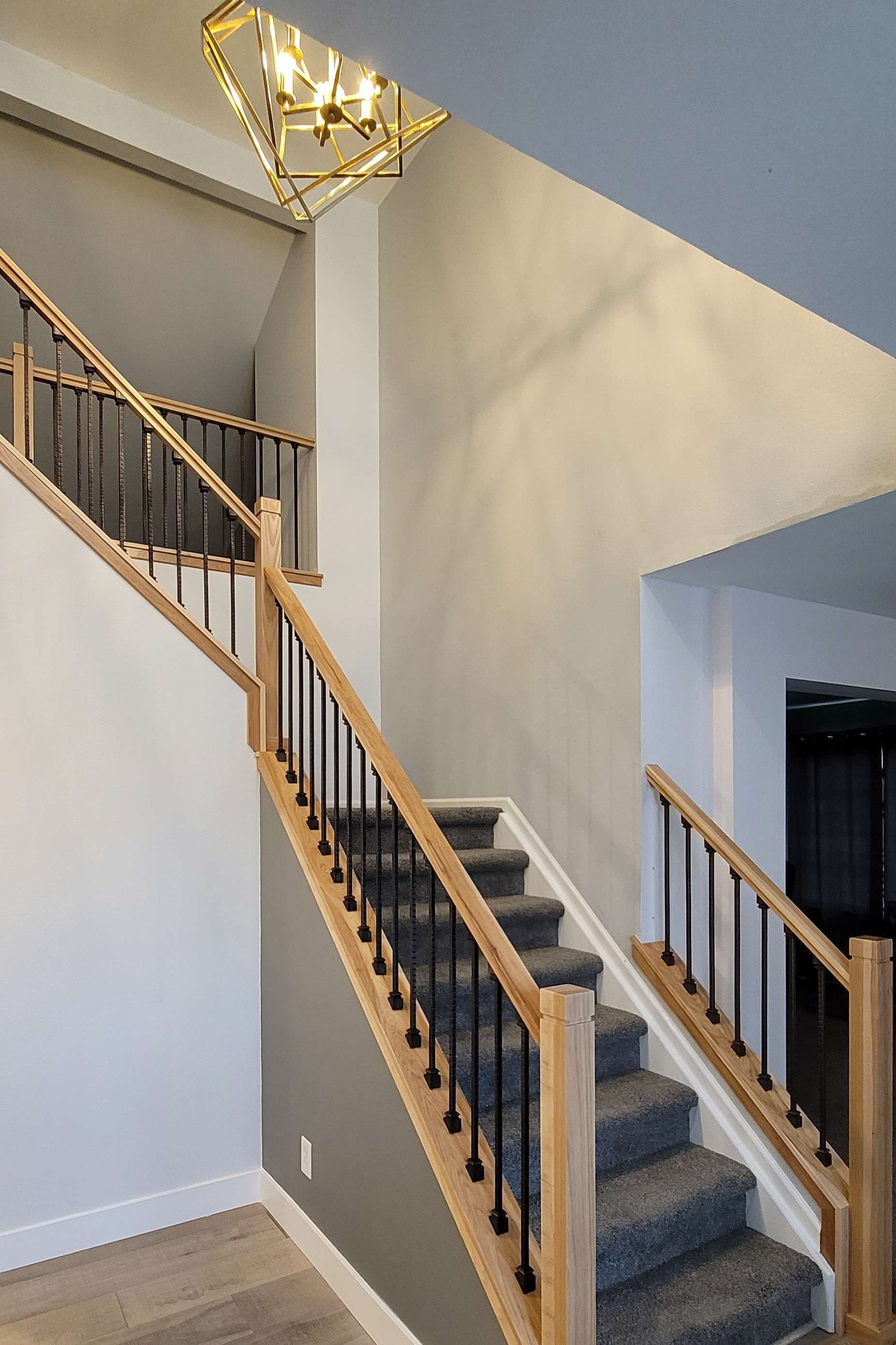 Staircase with light wood, black metal spindles, and gray carpet. A light fixture hangs overhead.