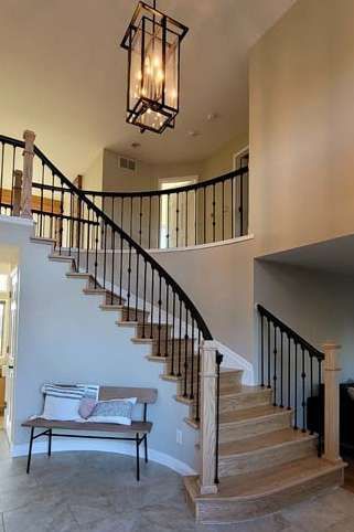 Elegant two-story foyer with curved staircase. Black metal railings, light wood stairs, chandelier, and a bench.