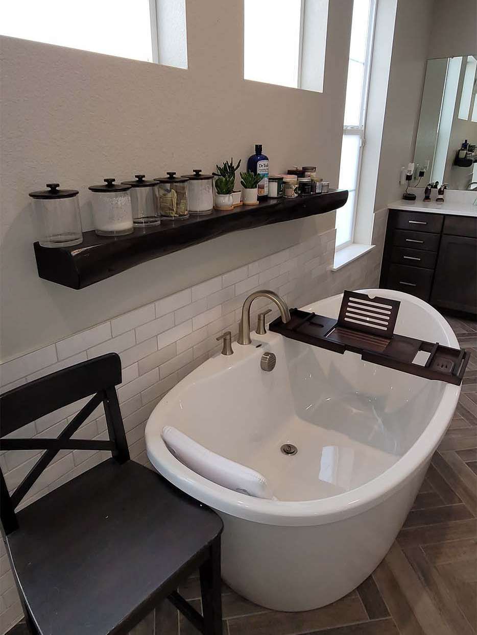 Bathroom with a white oval bathtub, dark wood shelf holding jars and plants, and a dark chair.