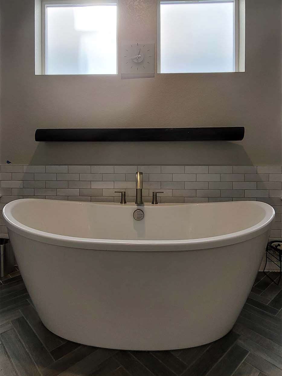 Freestanding white bathtub with brushed nickel fixtures, under windows, below a black shelf and tiled wall.