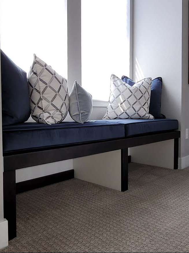 Window seat with navy cushions and patterned pillows against a bright window, on beige carpet.