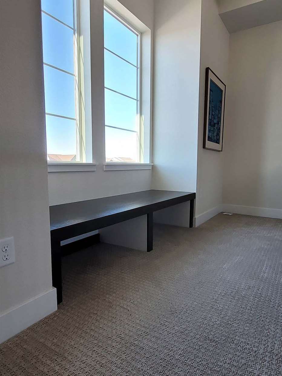 Window seat with black bench in a room with a window, white walls, and beige carpet. Blue artwork on wall.