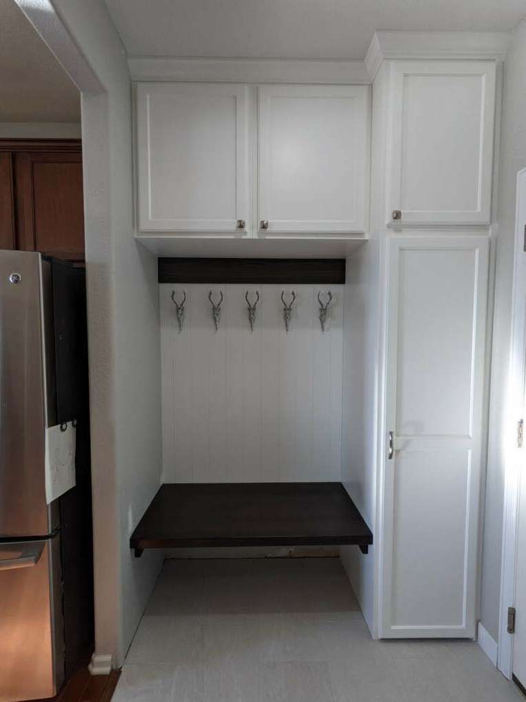 White built-in storage unit with cabinets, hooks, and a dark shelf. Located next to a refrigerator.