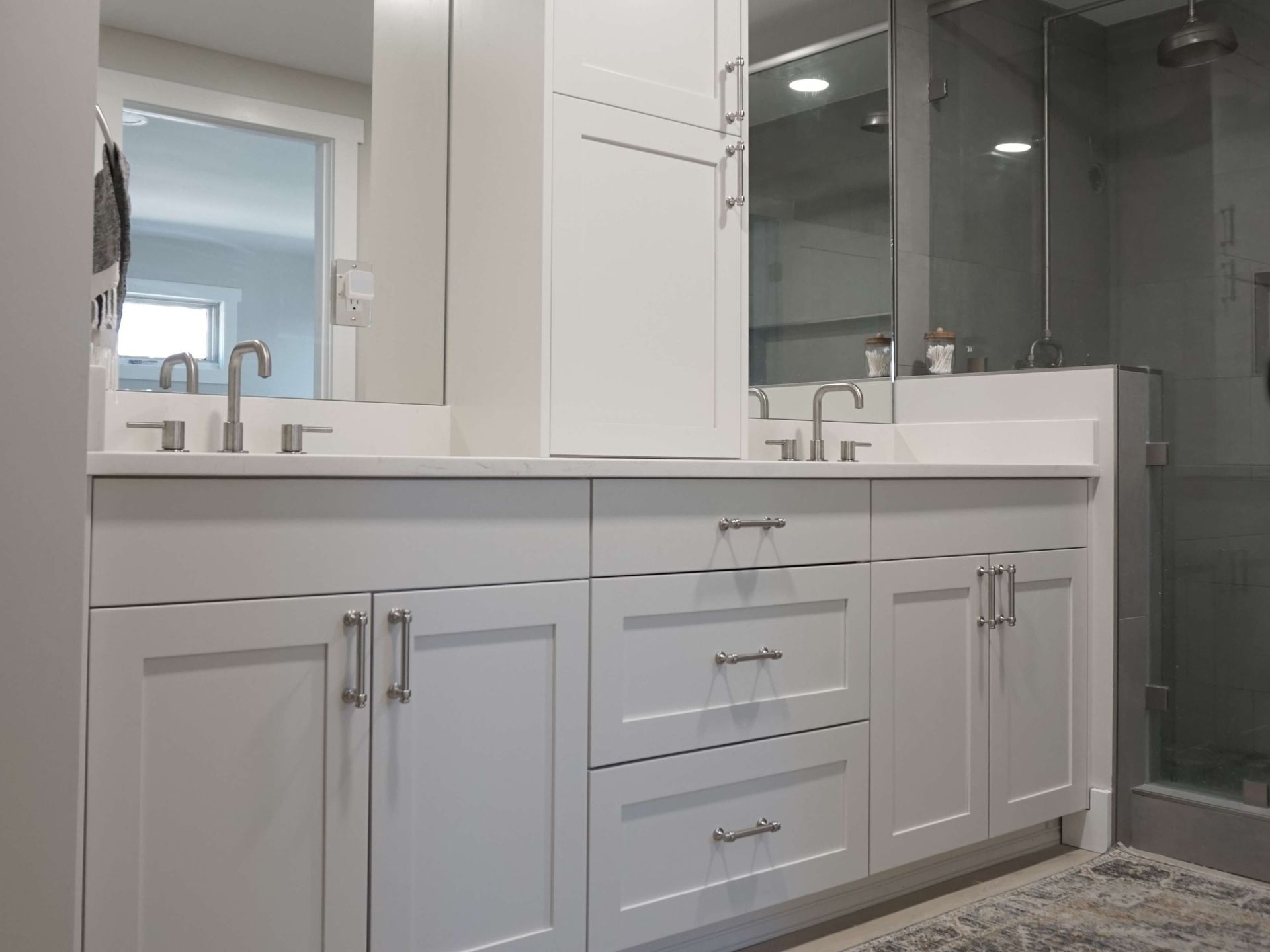 White bathroom vanity with double sinks, cabinets, and a mirror. A shower with glass doors is visible.