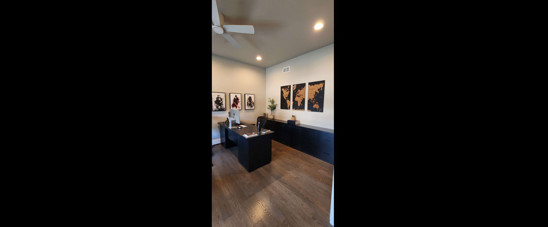 A well-lit office with dark furniture, wooden floor, and portraits on the wall. Ceiling fan visible.