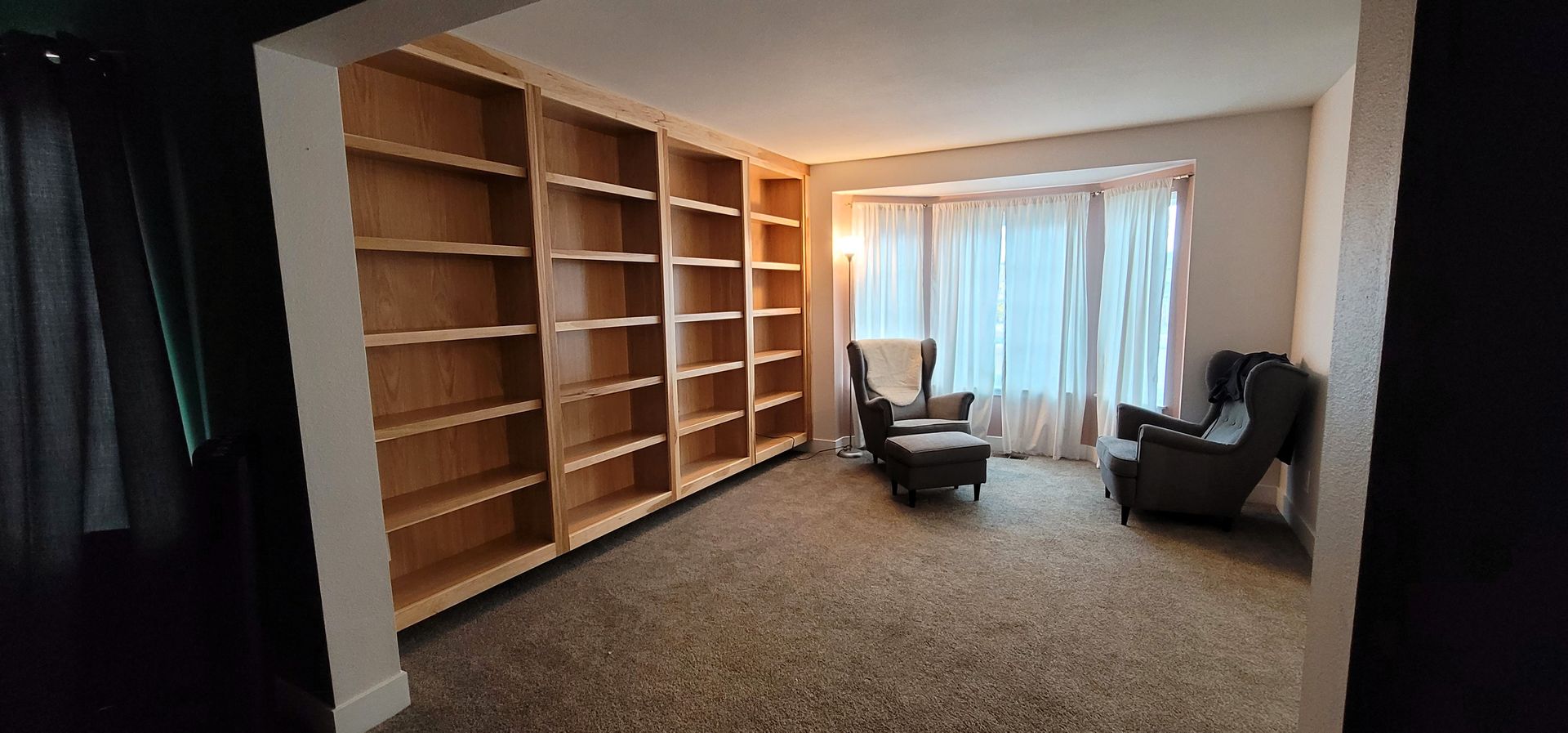 A room with built-in bookshelves, two armchairs, a footrest, and a window with sheer curtains.