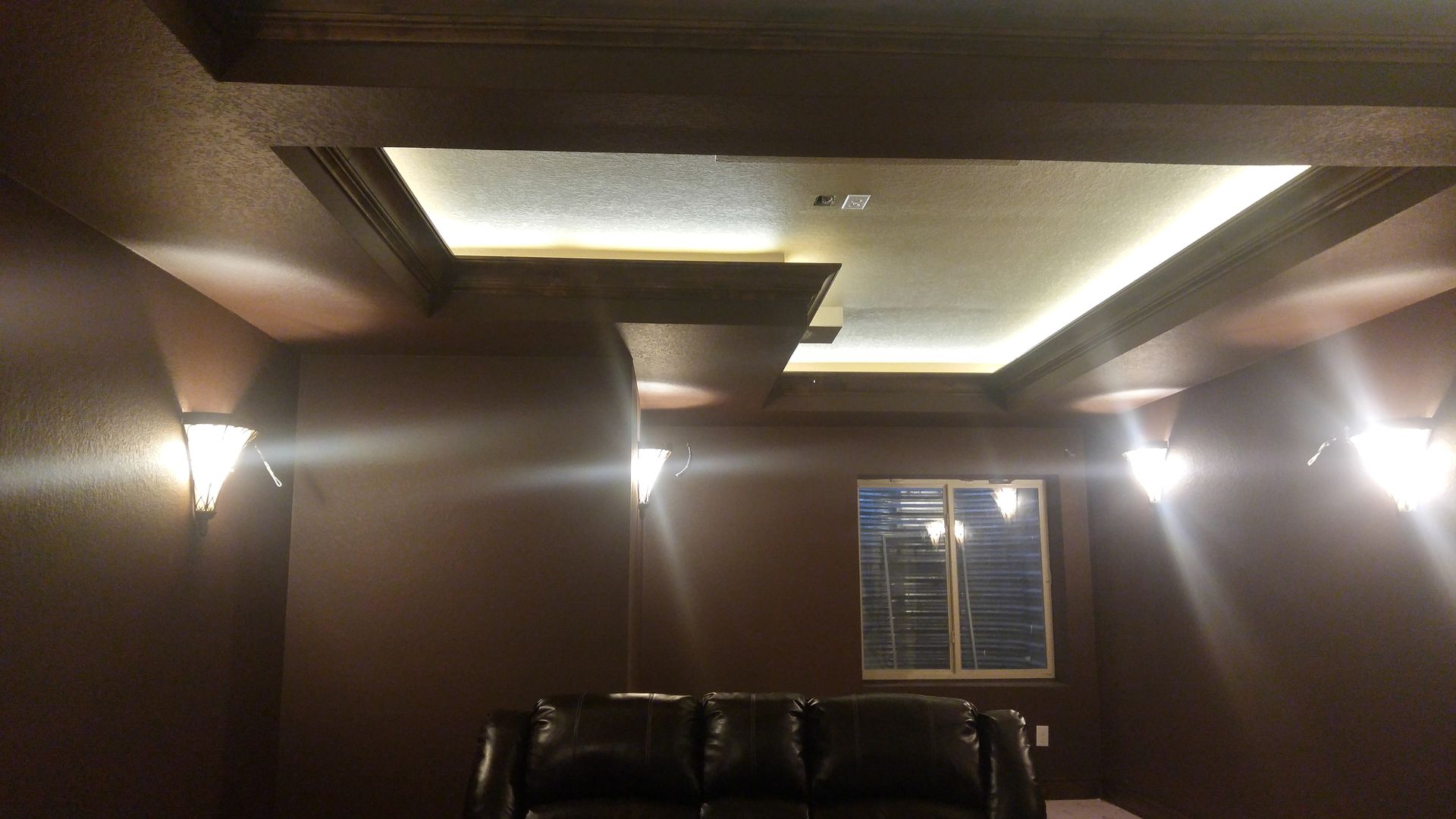 Dark brown room with illuminated ceiling beams, sconce lights, and a black couch.