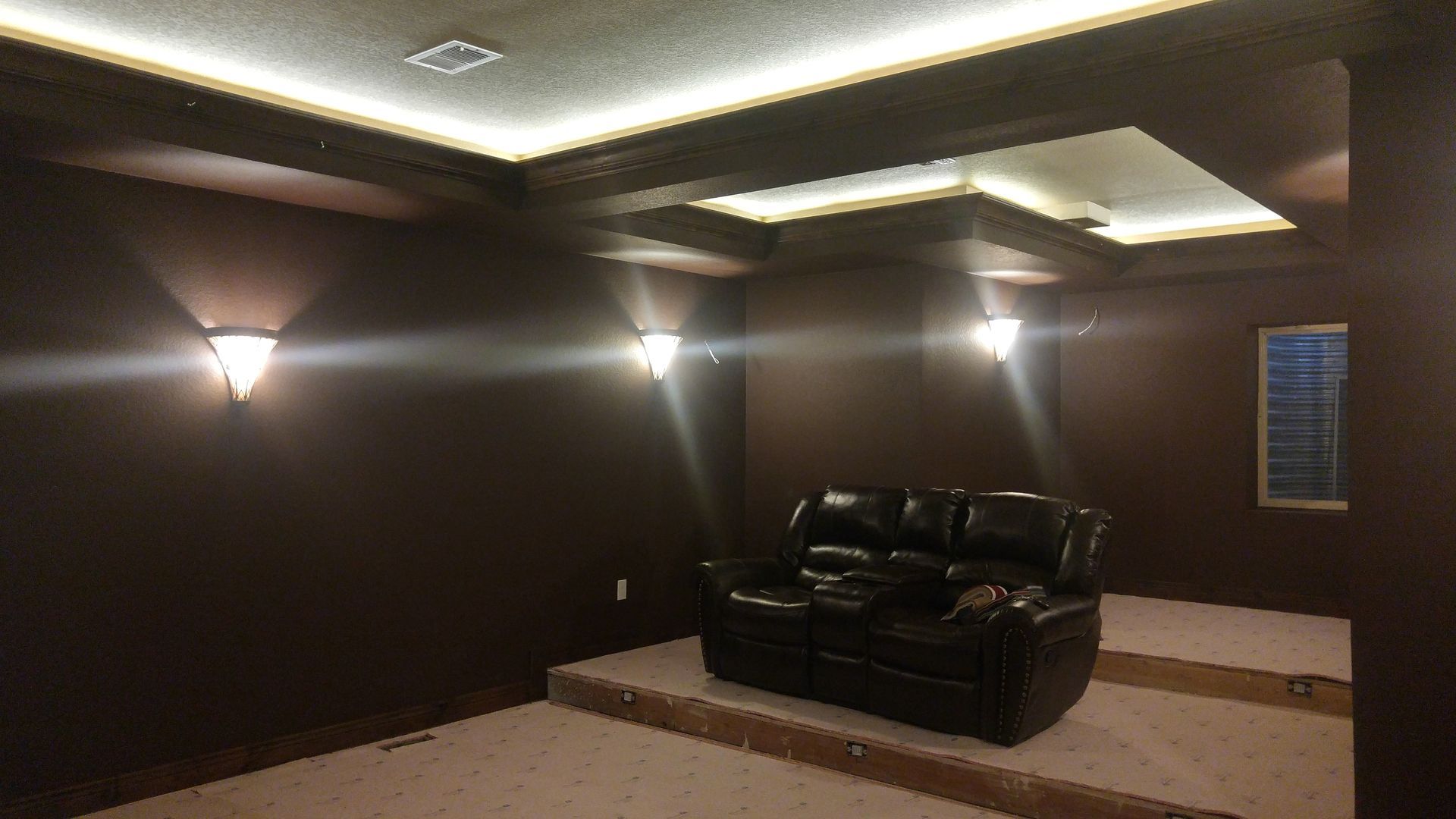 A dark home theater with a black leather recliner on a raised platform, wall sconces, and ambient ceiling lighting.