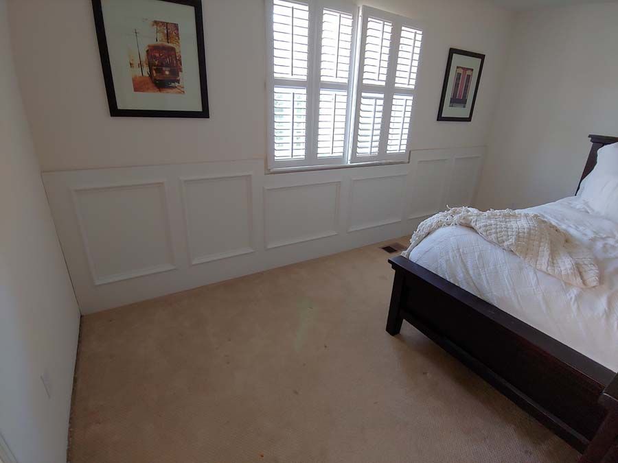 Bedroom with wainscoting, window with shutters, art, and a dark wooden bed with white bedding. Beige carpet.