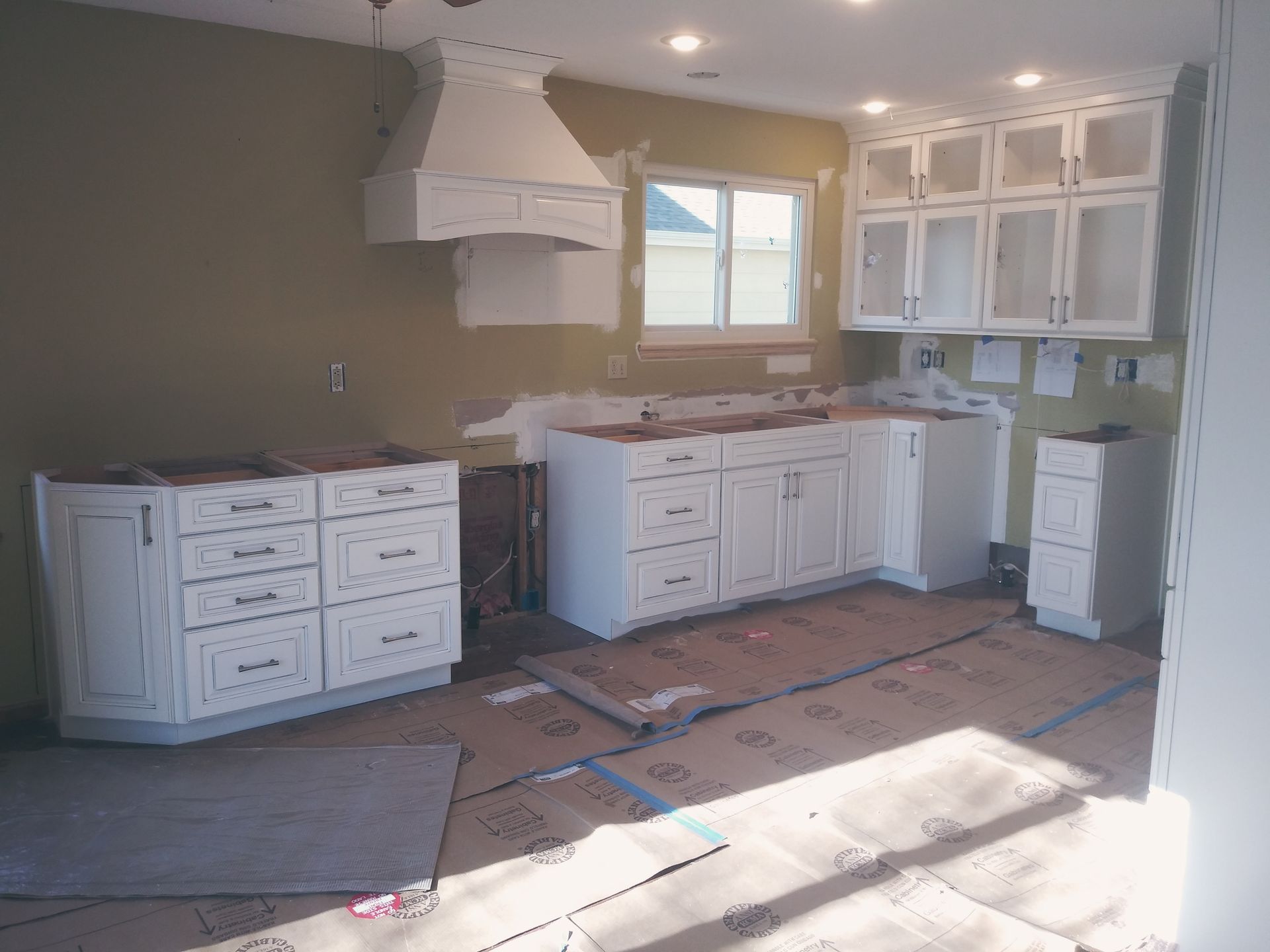 Kitchen under construction with white cabinets, hood vent, and window, unfinished walls, and flooring.