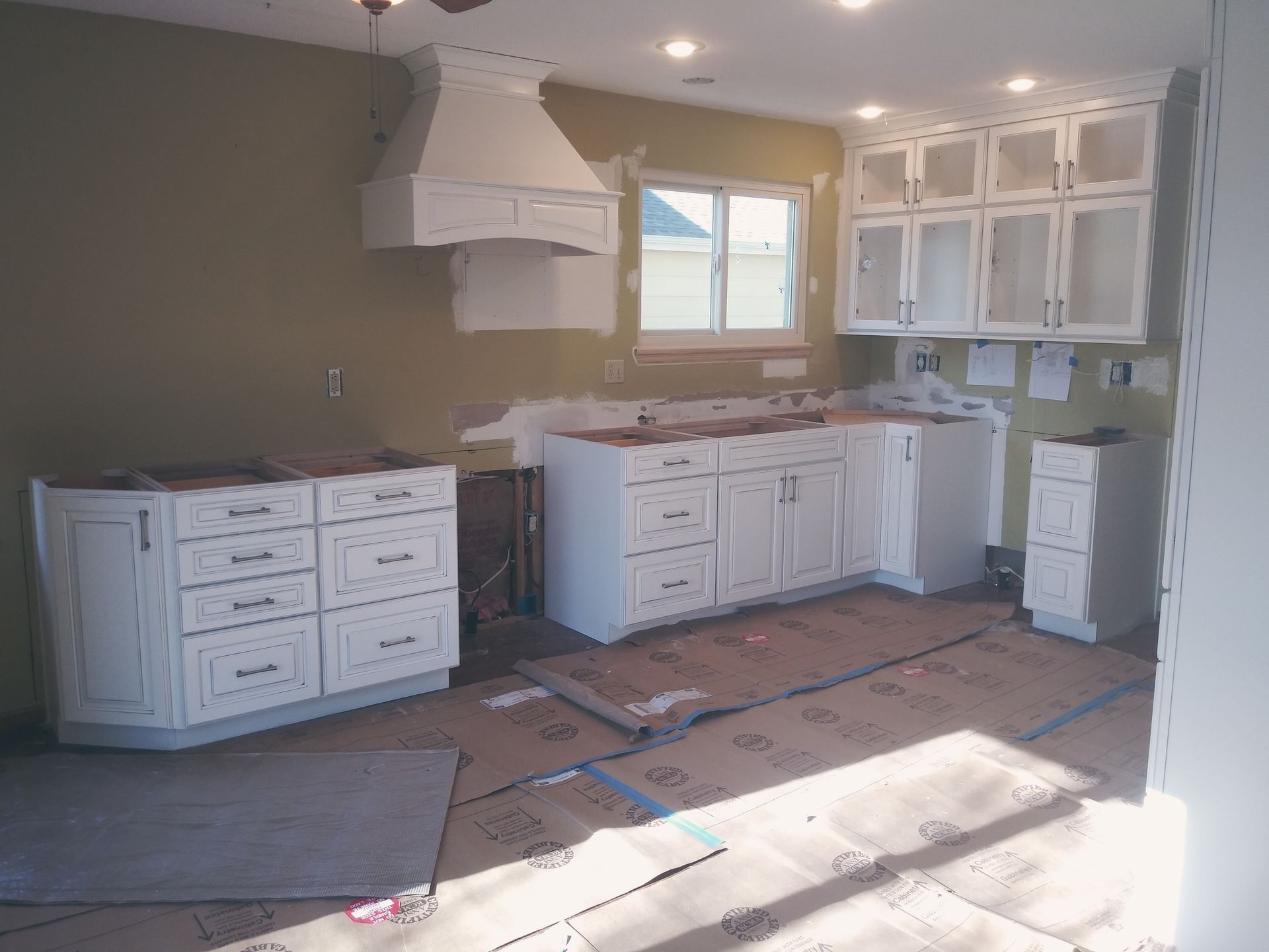 Kitchen renovation with white cabinets, range hood, window, and unfinished walls.