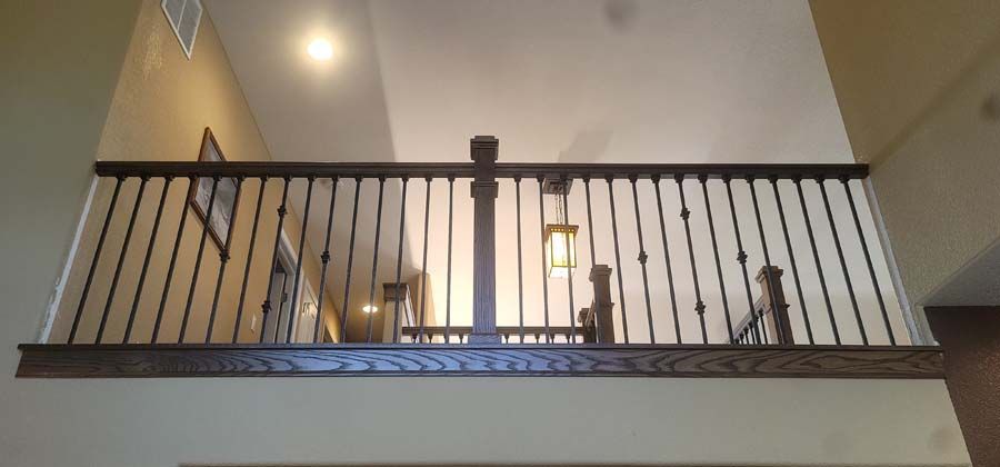 A wooden railing with black iron spindles on an elevated interior balcony with a high ceiling and a hanging light fixture.
