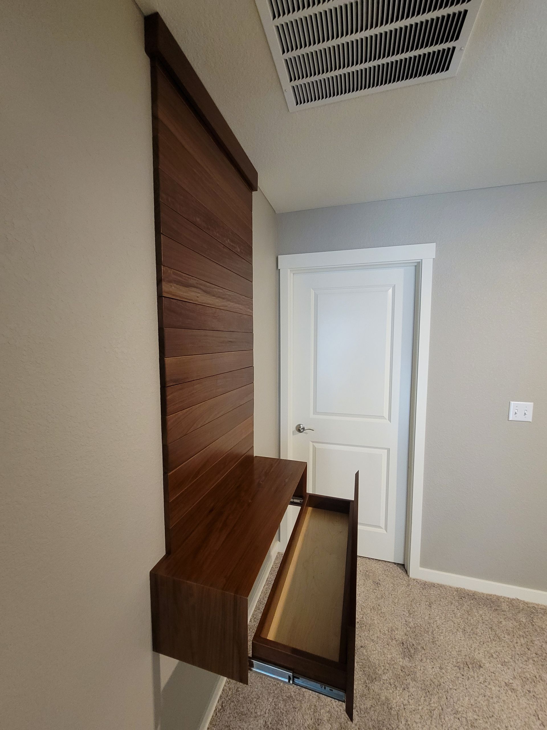 Wall-mounted wooden desk with an open drawer, attached to a textured wall. A door and vent are in the background.