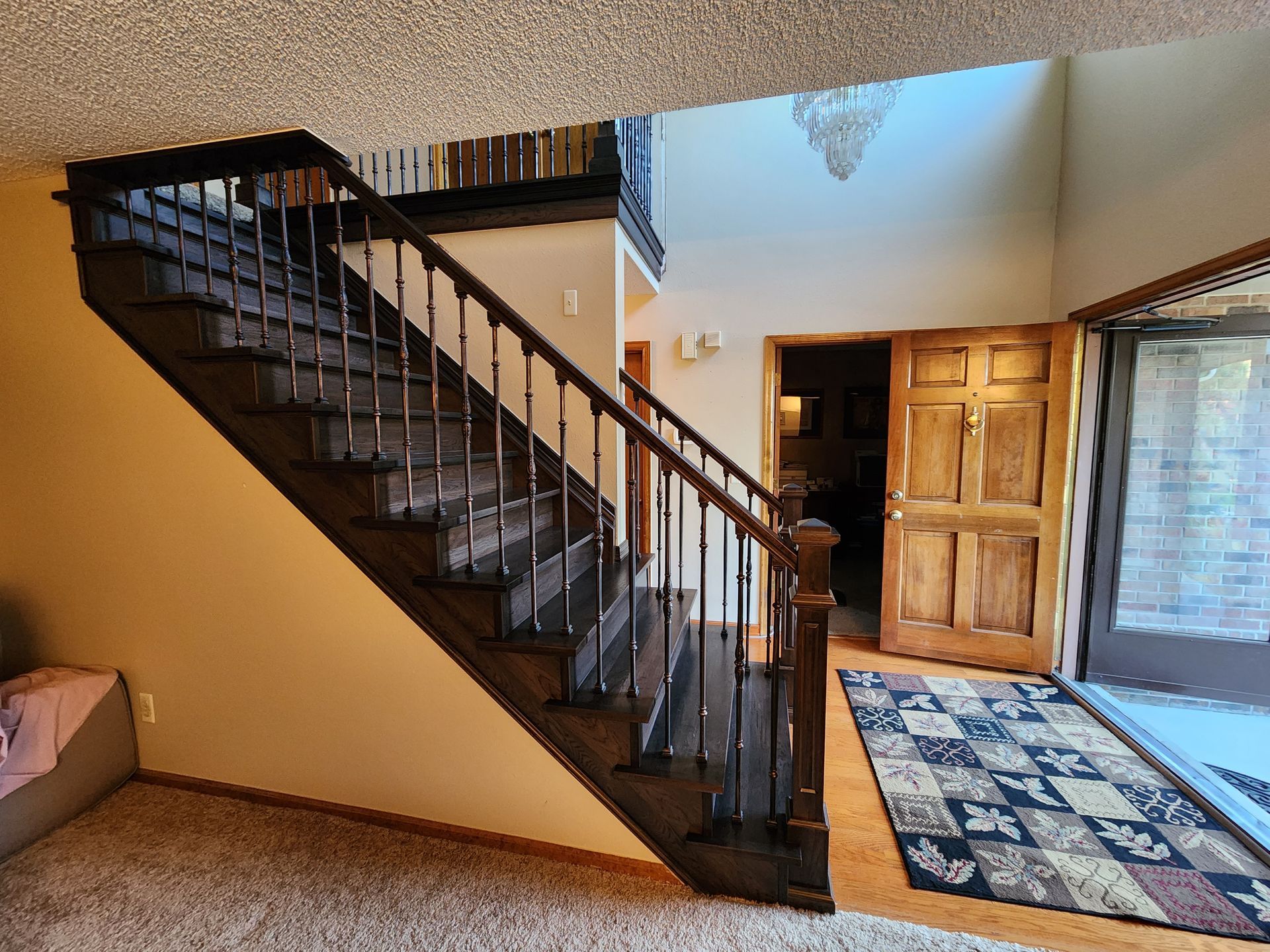Interior view of a staircase with dark brown wooden steps and railings. A wooden door and rug are visible.