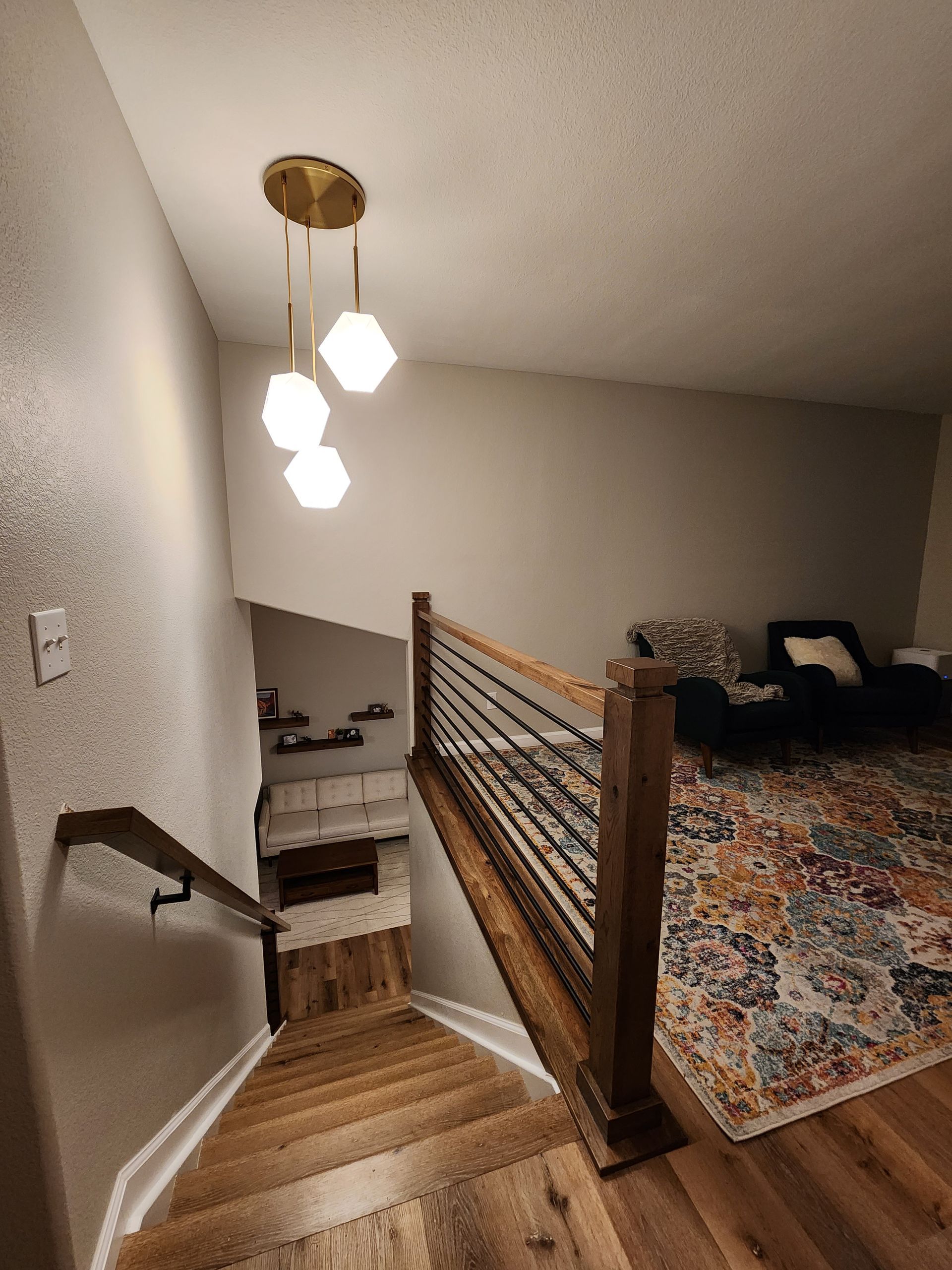 Staircase with wooden steps and railing, leading down to a living room with a rug and seating. Modern light fixture.