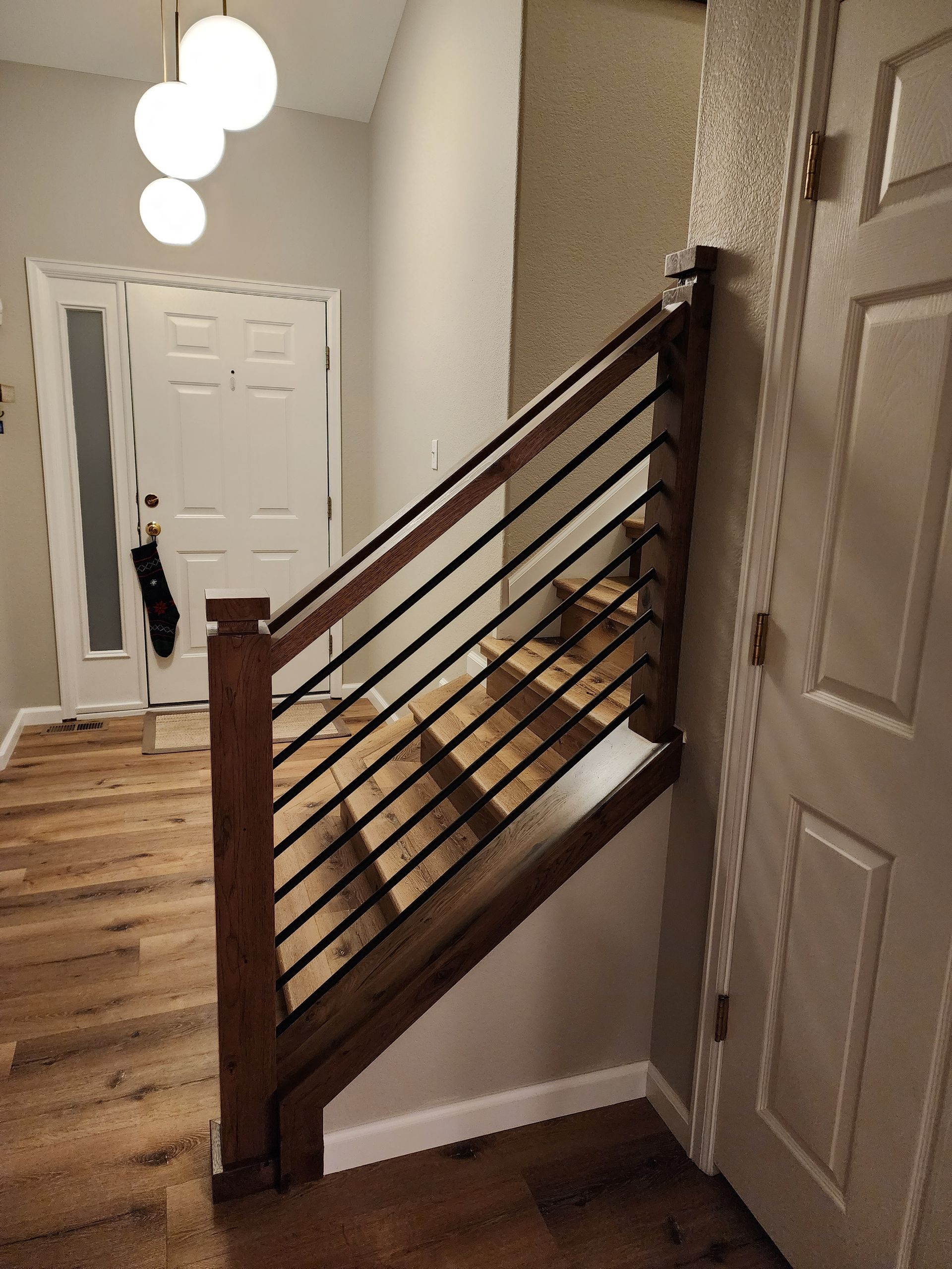 Staircase with wooden steps and railing, black horizontal bars. White door, pendant lights, wooden floors.