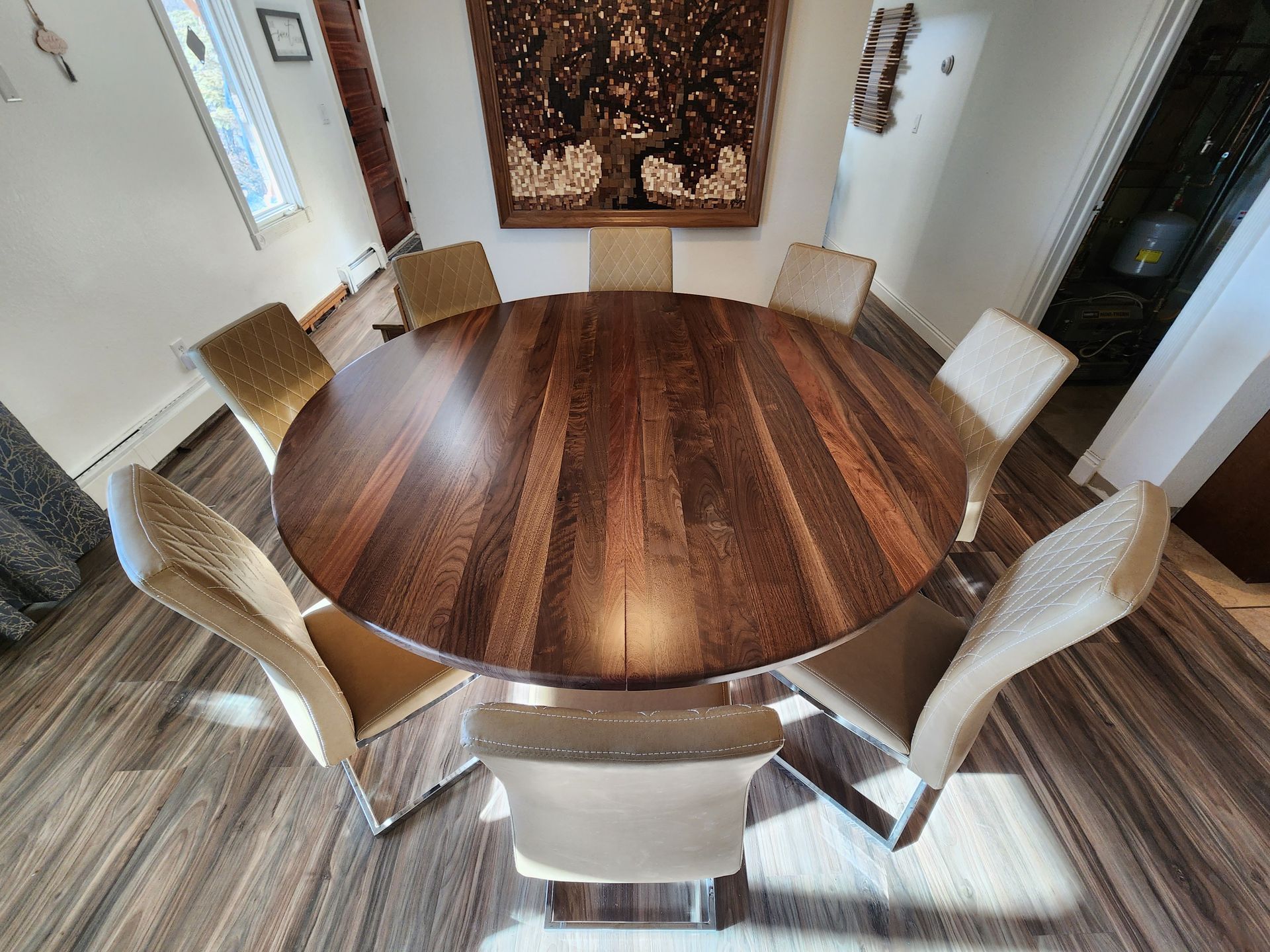 Round wooden dining table with eight beige chairs in a sunlit room.