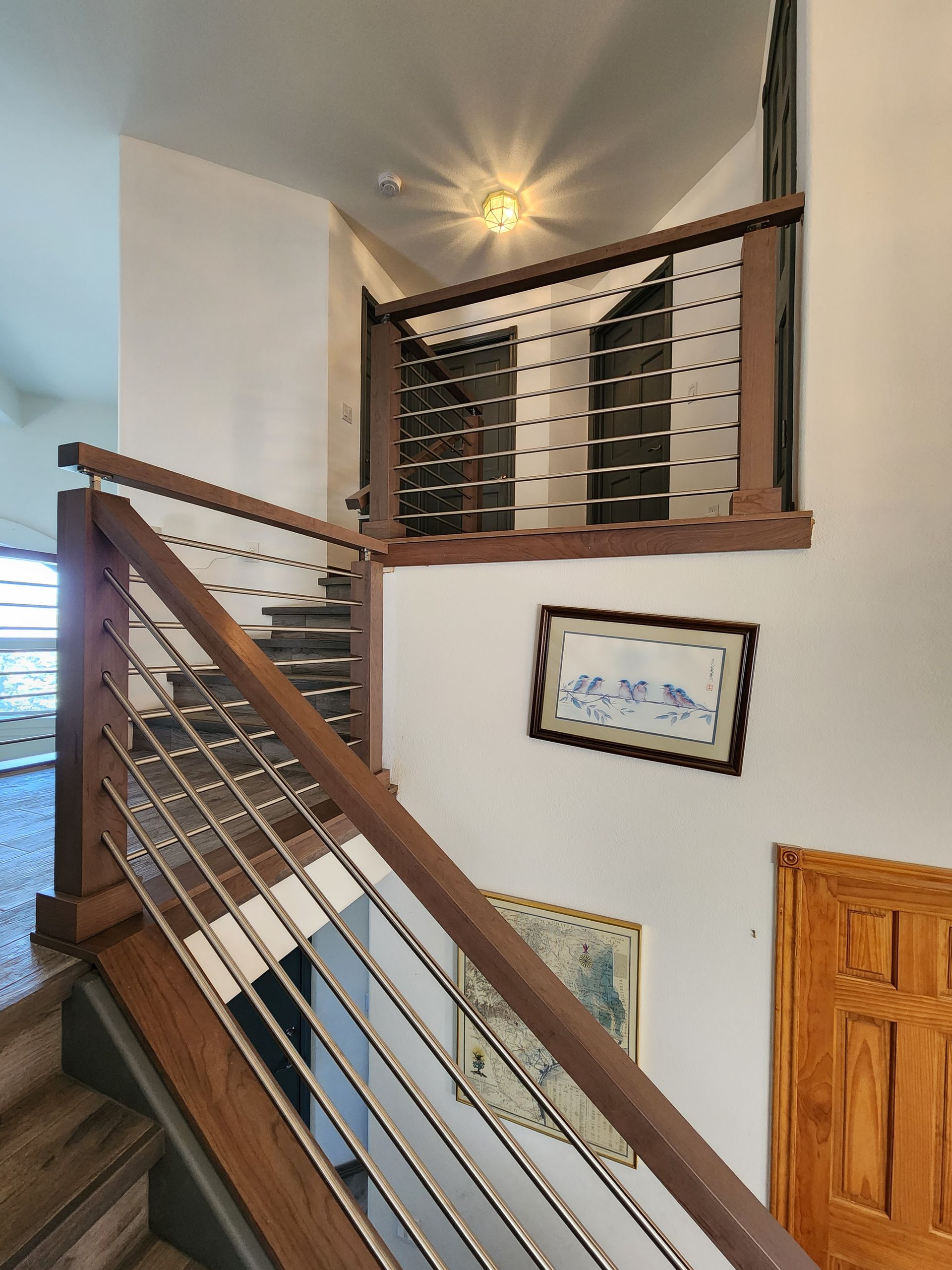 Wooden staircase with metal cable railings, leading to a second floor. Artwork hangs on the wall.