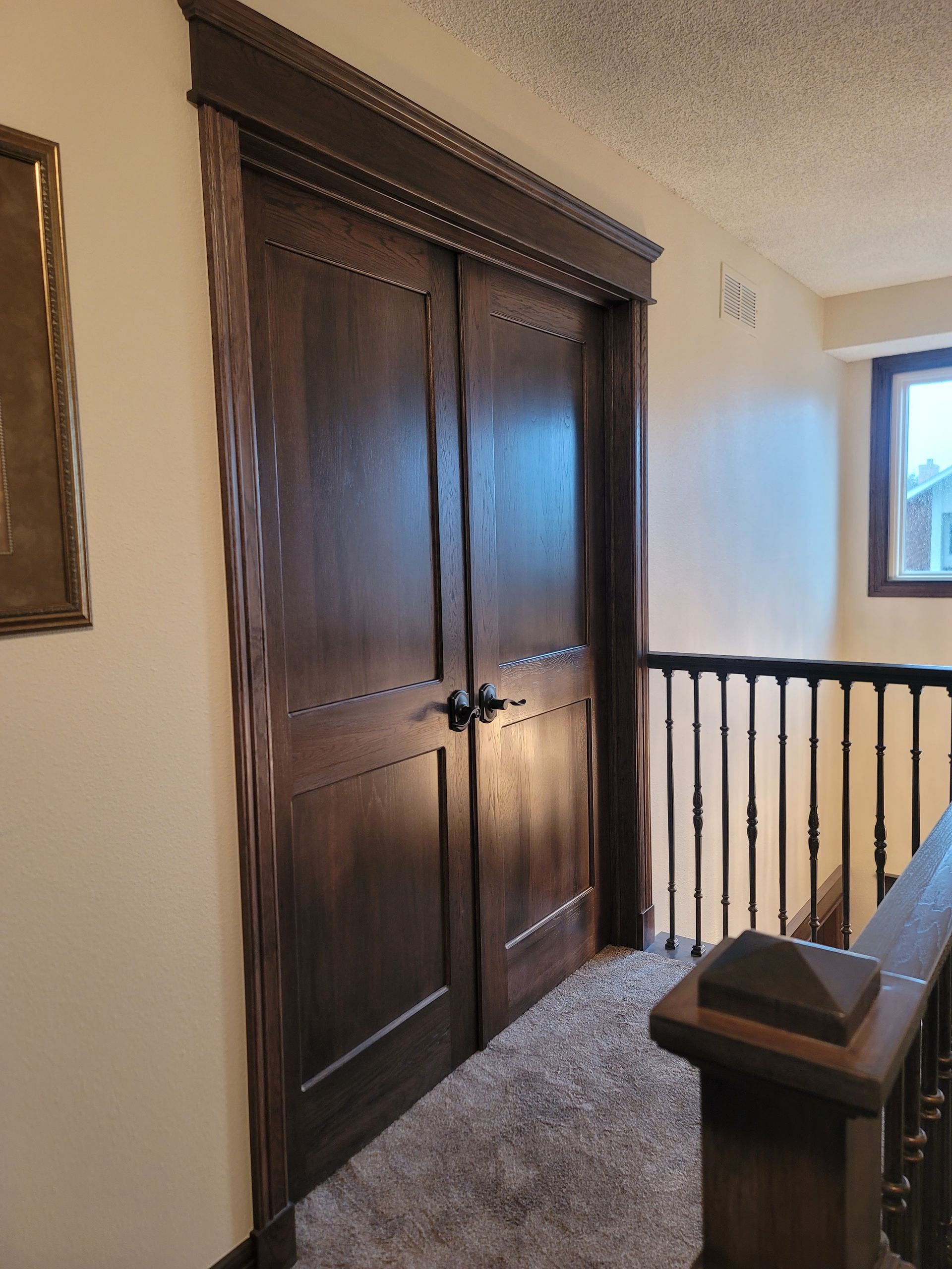 Dark wood double doors with detailed trim, in a hallway with carpet and railing.