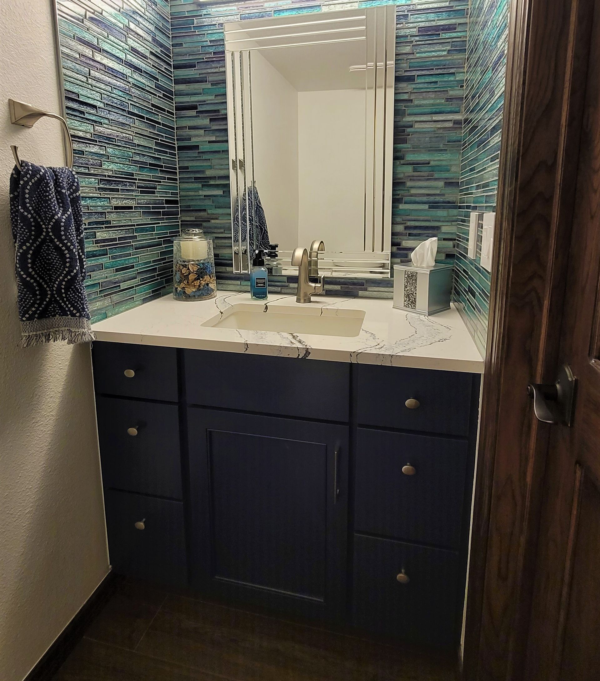 Bathroom with blue-tiled walls, navy vanity, white countertop, silver faucet, and decorative mirror.