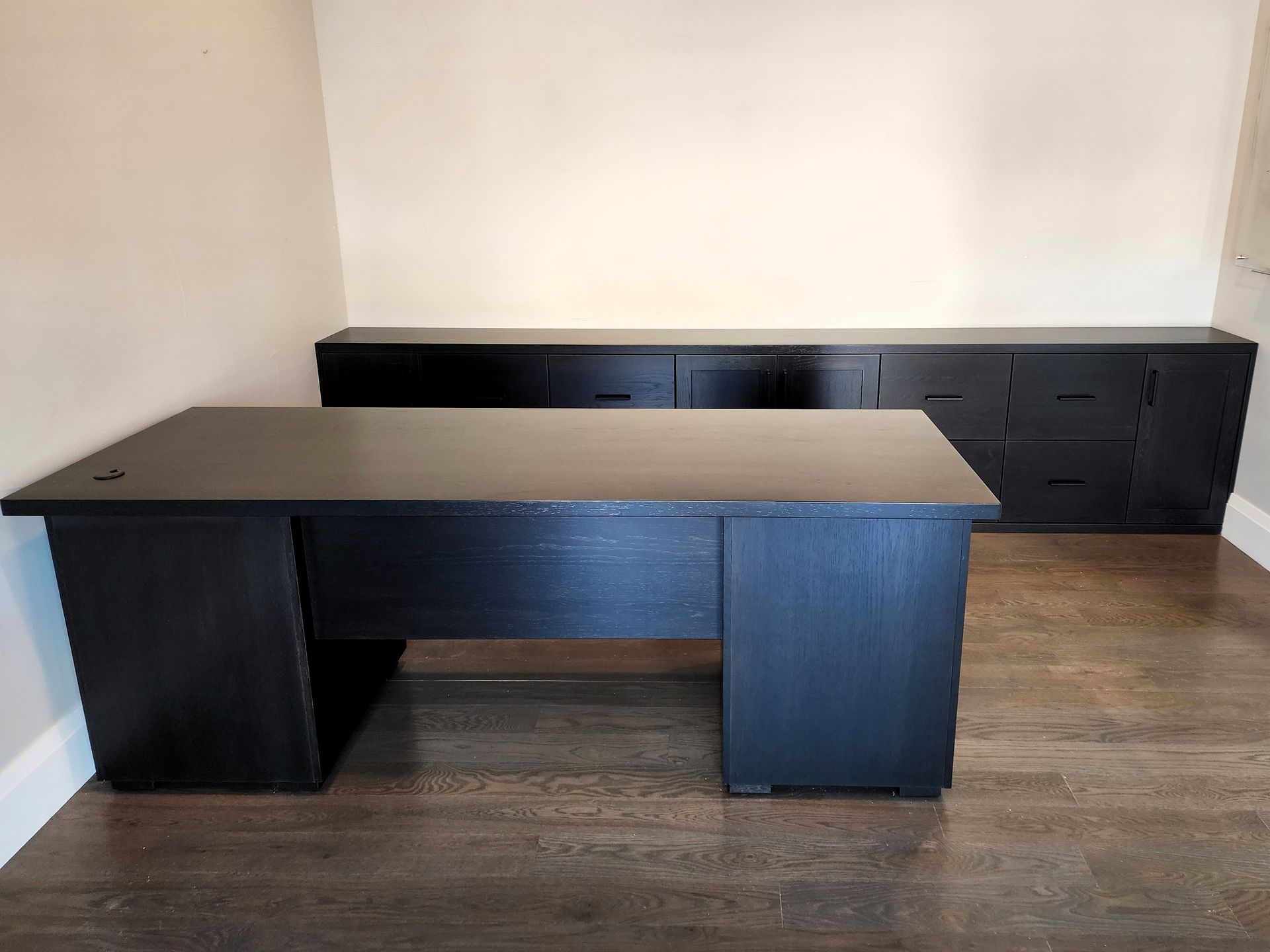 Black desk and matching storage cabinet in an office space with wood flooring and cream walls.