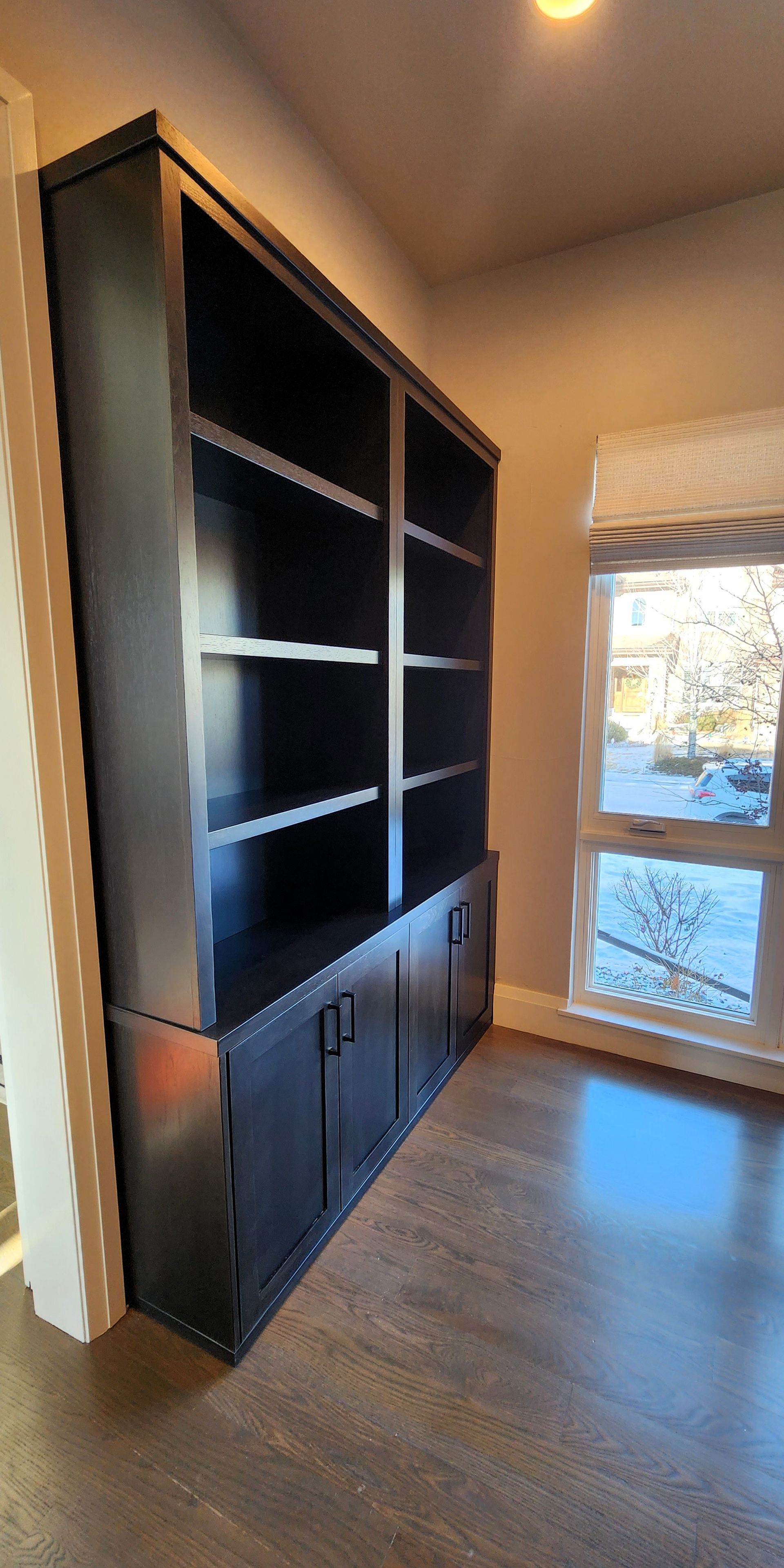 Black built-in bookshelf with cabinet doors, set against a wall. Wooden floors and a window visible in the background.