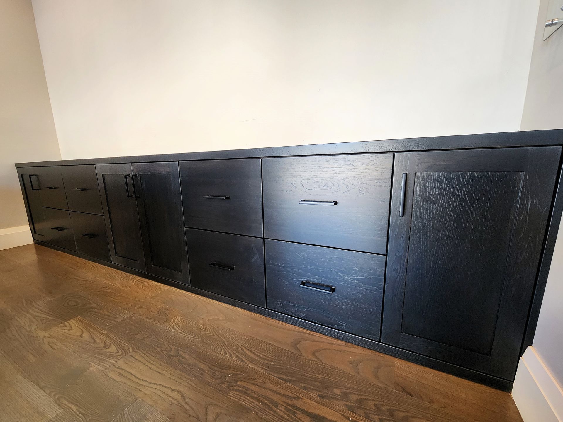 Black cabinet with doors and drawers against a white wall and hardwood floor.