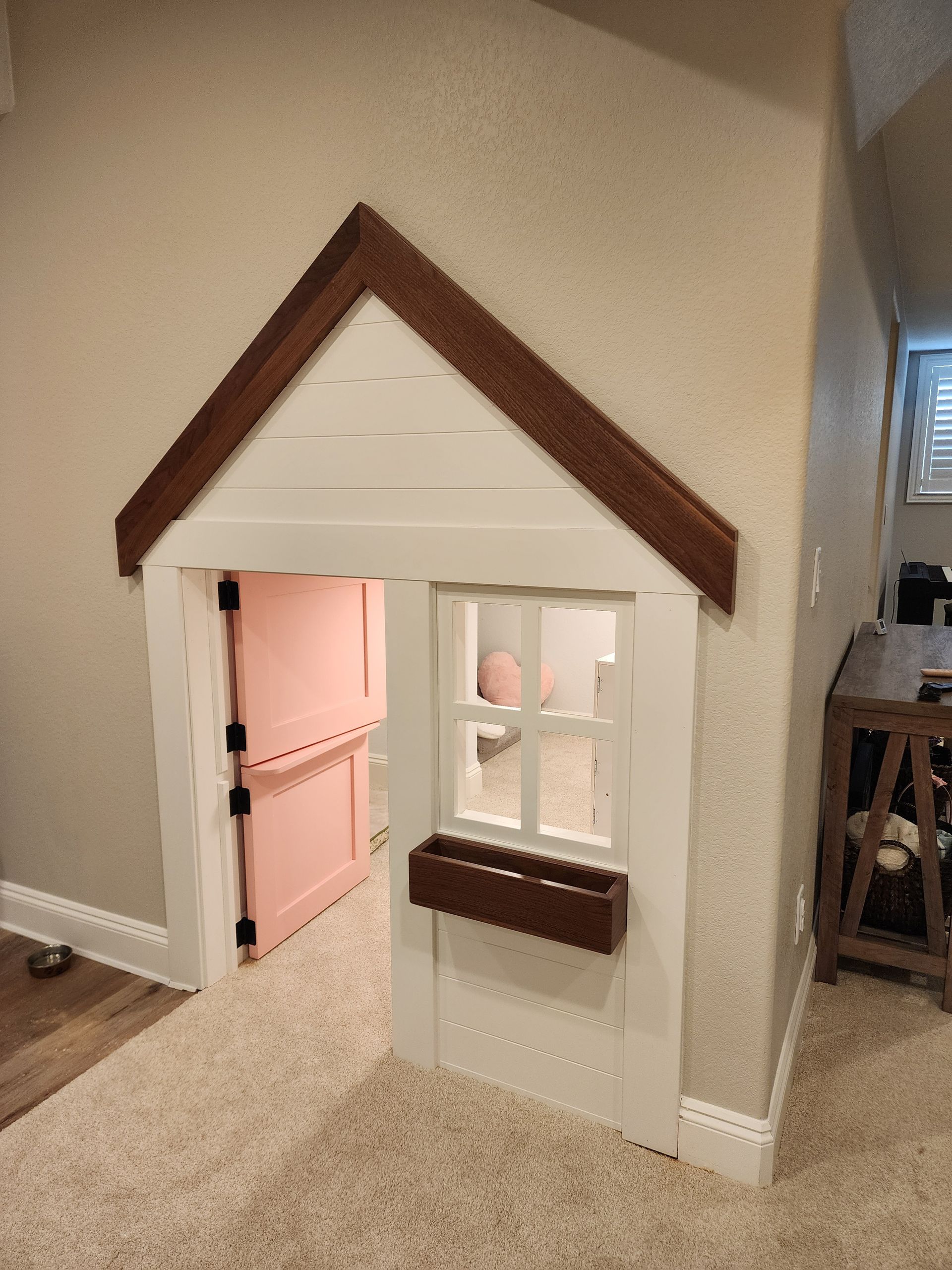 Playhouse built into a wall, with a door, window, and brown roof trim. Pink door opens into the playhouse.