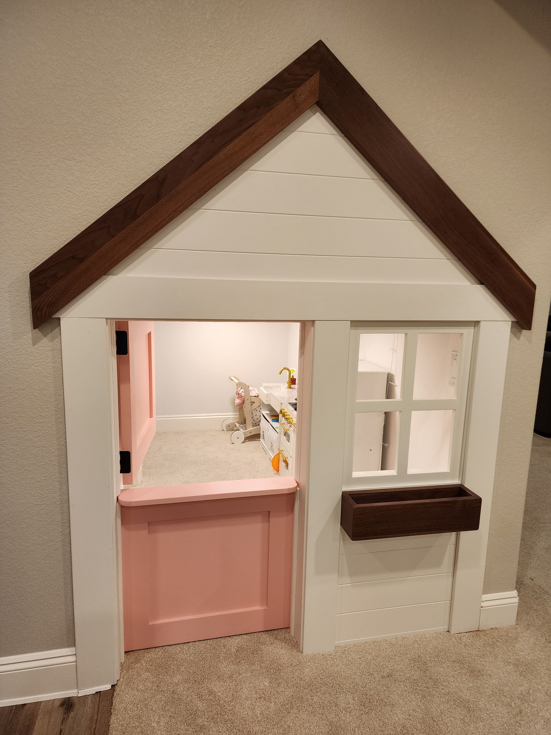 Child's playhouse built into a wall with pink door and window, white exterior, brown trim.