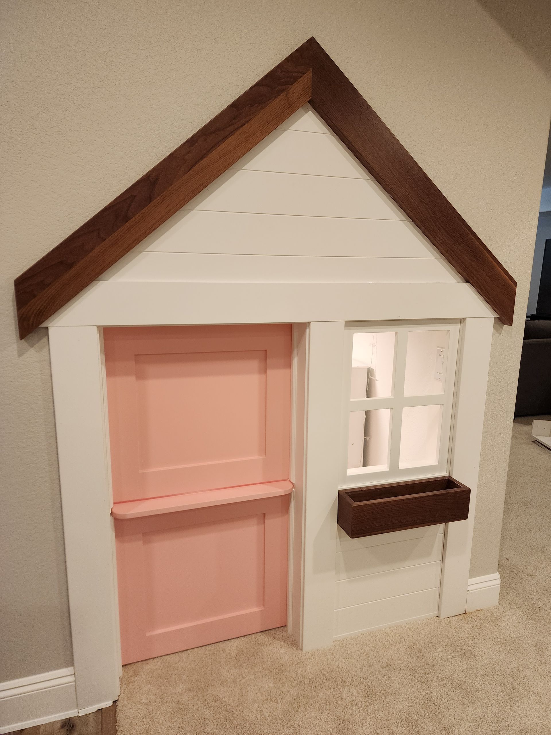 Playhouse built into a wall, with pink door, white siding, brown window box, and a brown roof.