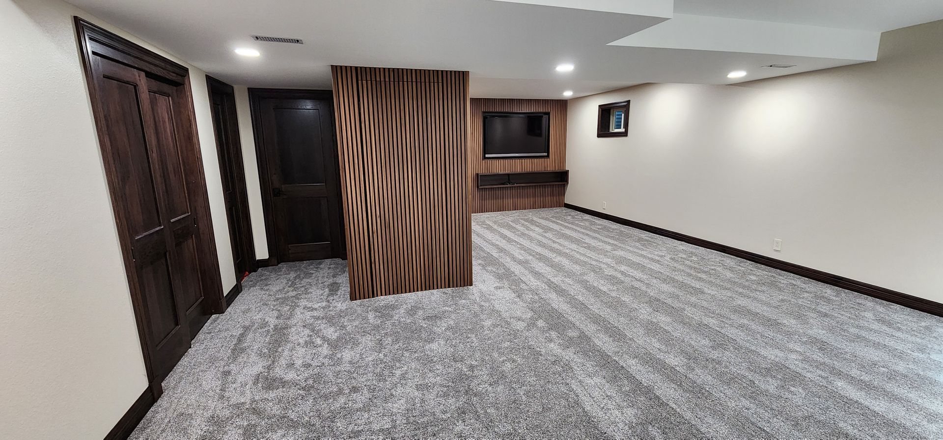 Room with gray carpet, a dark brown door, a wood-paneled wall, and a TV on a wall.
