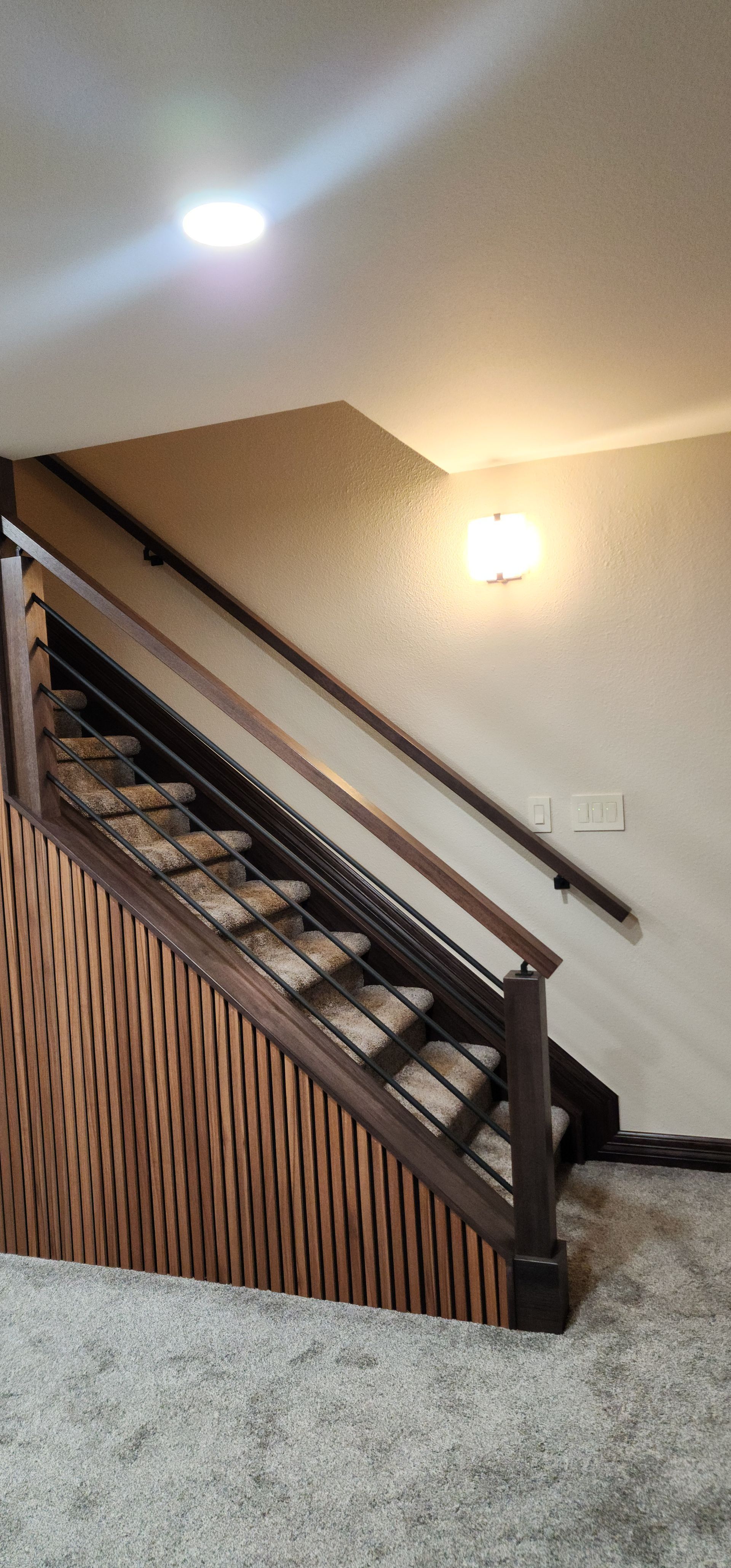 Wooden staircase with brown handrails and patterned side paneling, leading upwards. Carpet flooring.
