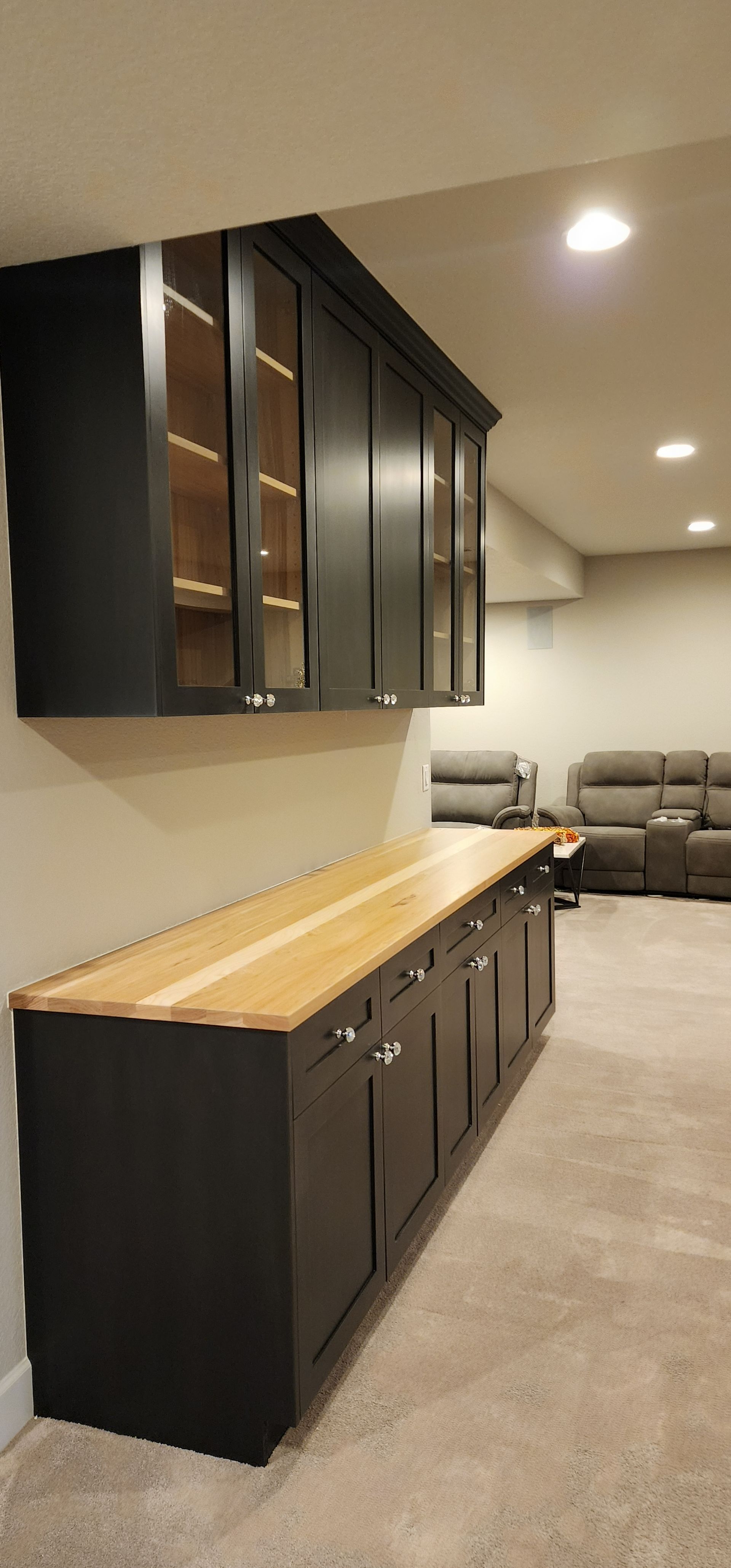 Dark cabinets with glass doors over a dark cabinet bar with a wood countertop. Gray carpet and wall.