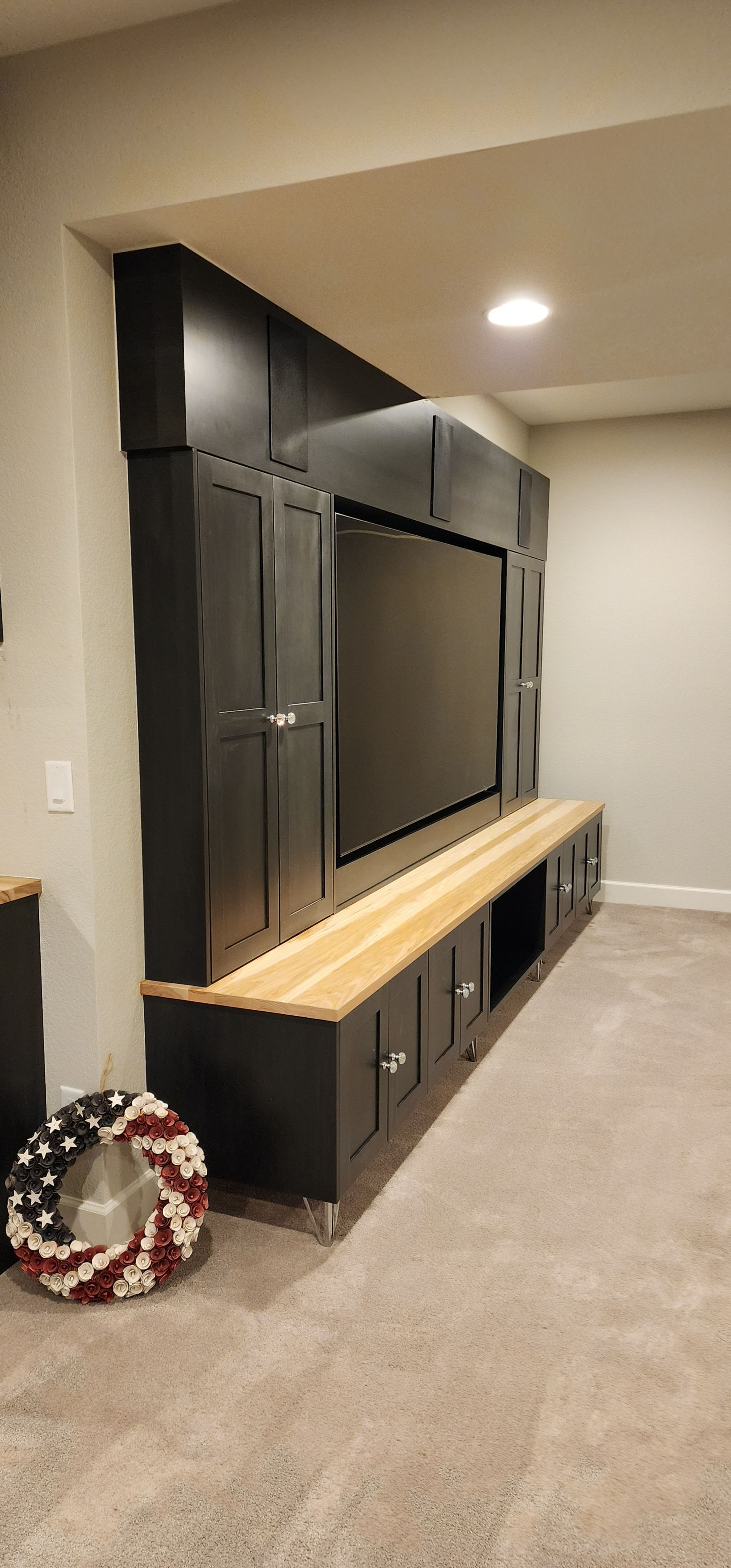 Built-in dark gray entertainment center with wooden bench and television in a room with light gray carpet.