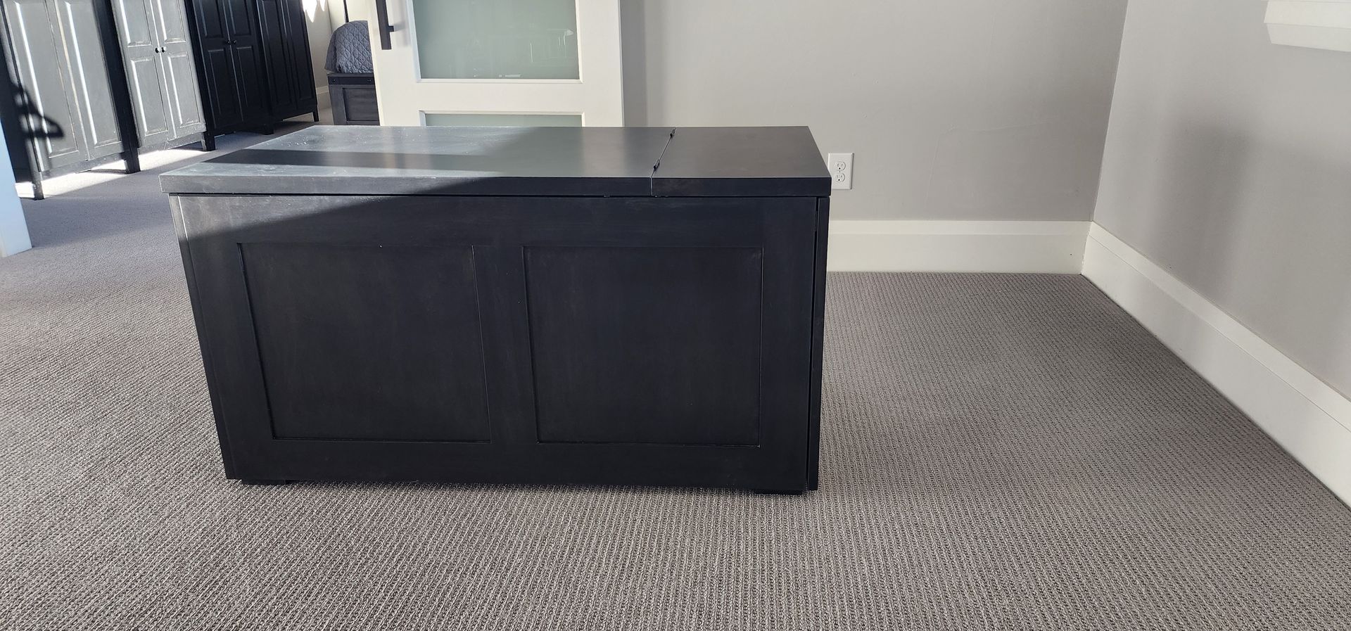Black storage chest on patterned carpet, beside a gray wall with white trim.