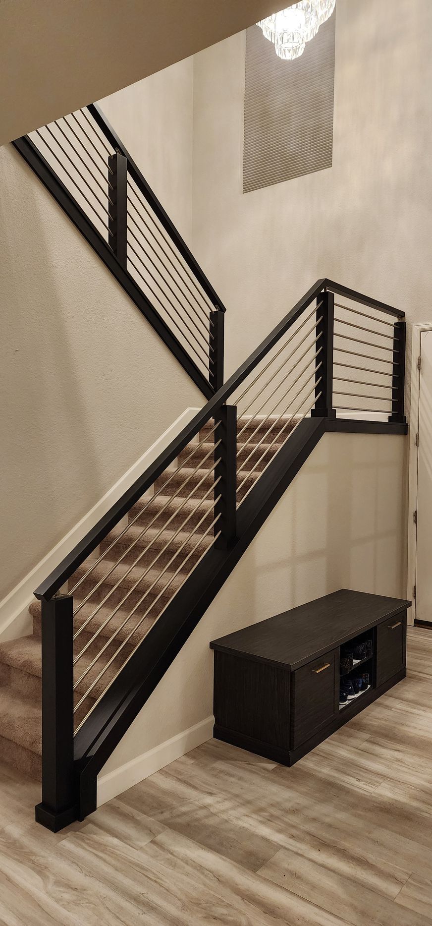 Staircase with black railing and horizontal metal cables, beige walls, and wood-look flooring. A dark cabinet sits below.