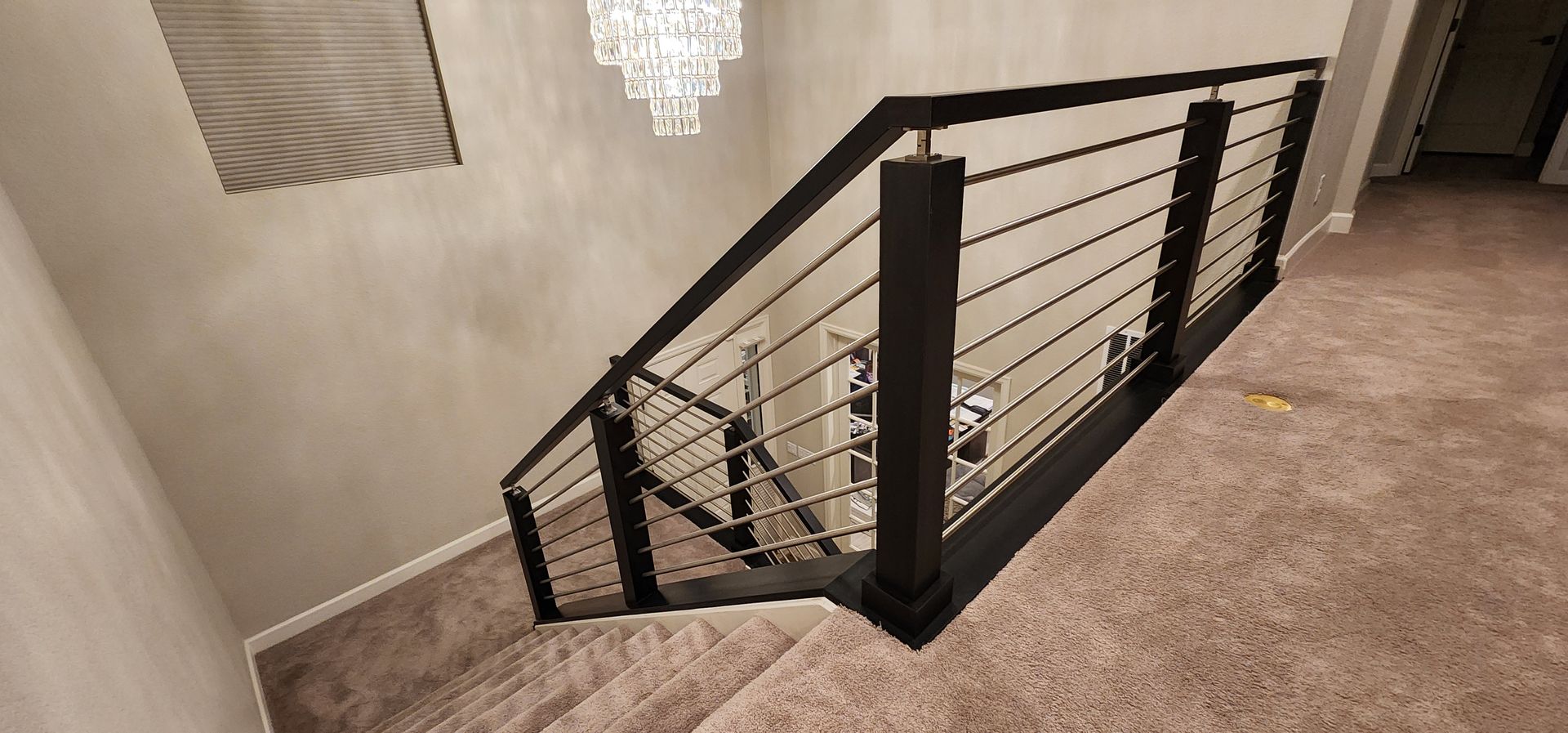 Modern staircase with black railing and cable infill, tan carpet, and chandelier.