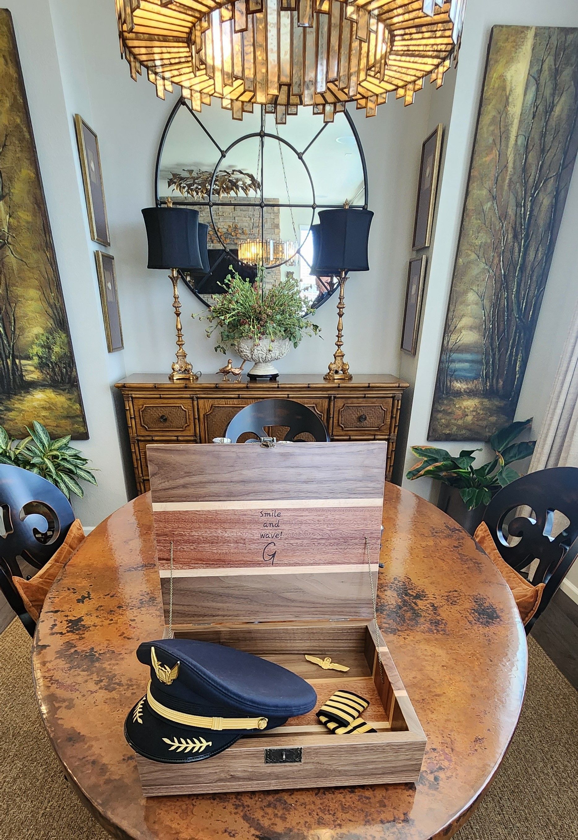 Pilot hat and pins in a wooden display on a round table, ornate mirror and lamps in background.