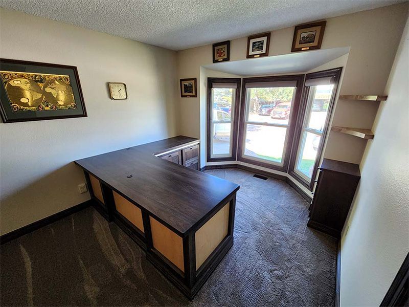 L-shaped dark wood desk in home office with a bay window and dark carpet.