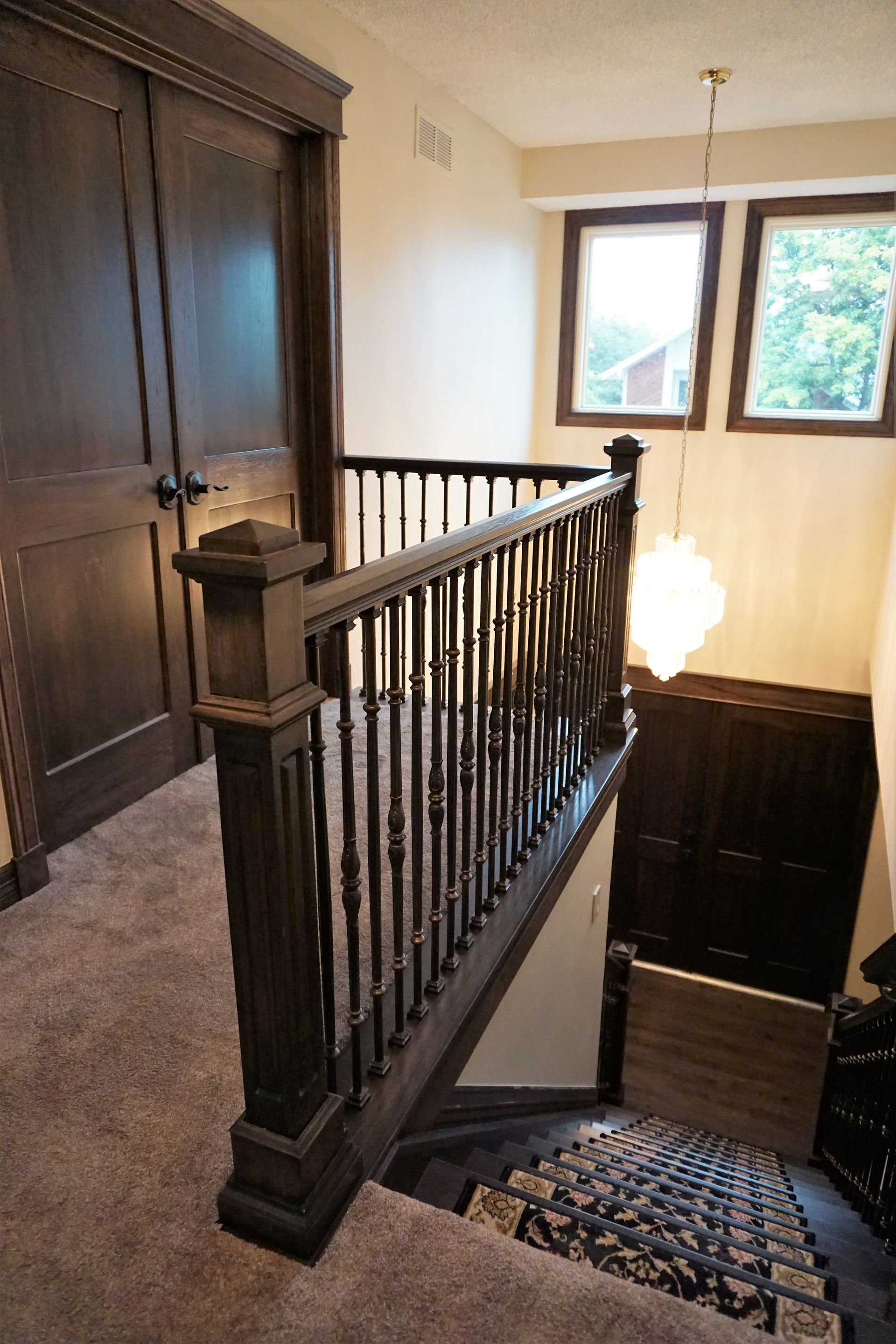 Dark wooden banister and staircase with carpet; two windows and a chandelier overhead.