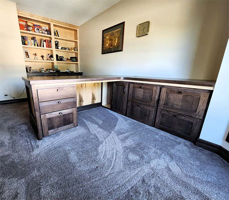 L-shaped wooden desk with drawers and bookshelves against a beige wall. Carpeted floor.