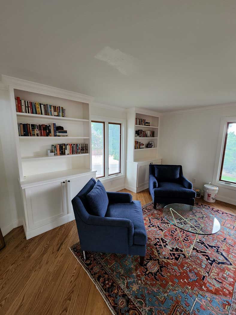 Living room with built-in bookshelves, blue seating, and a patterned rug. Sunlight streams through the windows.
