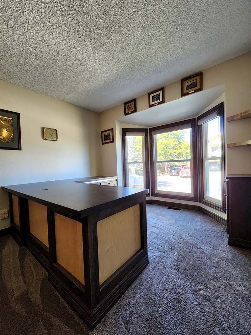 Dark desk in a room with a curved bay window. Carpet, framed art, and neutral walls.