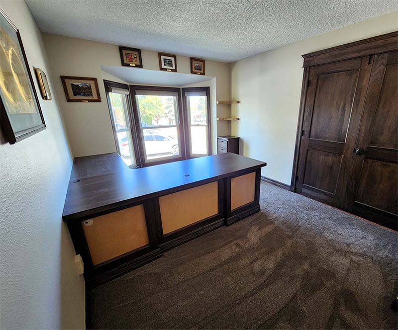 Office with built-in dark wood desk, cabinets, and bay window. Tan walls, brown carpet, and small framed pictures.
