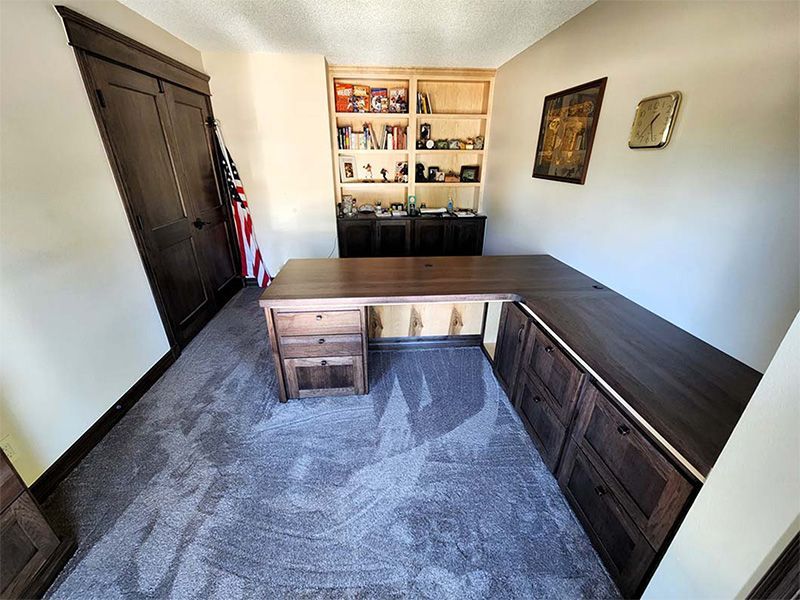 L-shaped wooden desk in office with built-in storage, bookshelves, American flag, and carpet.