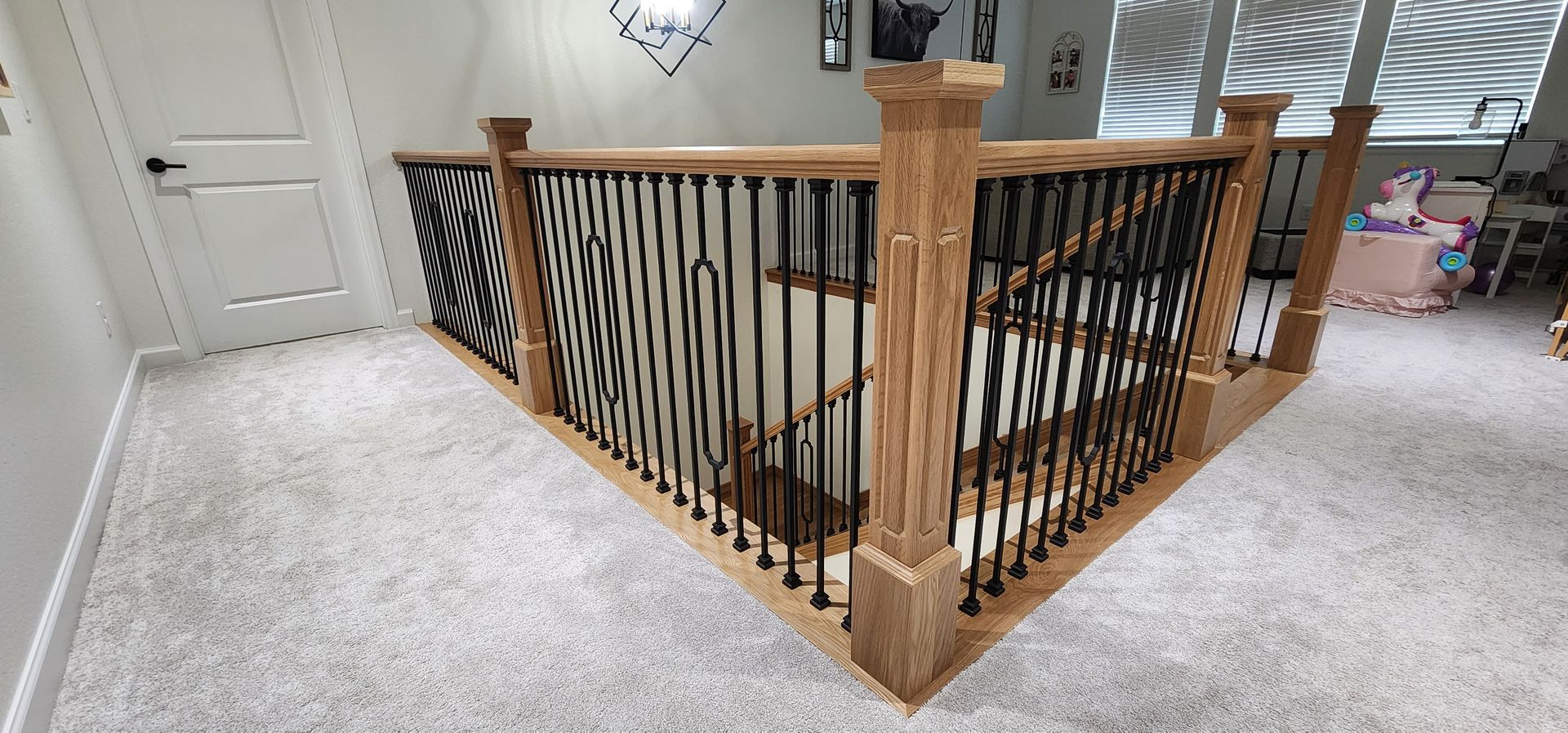Staircase with wooden railing and black spindles, viewed from above. Light gray carpet.