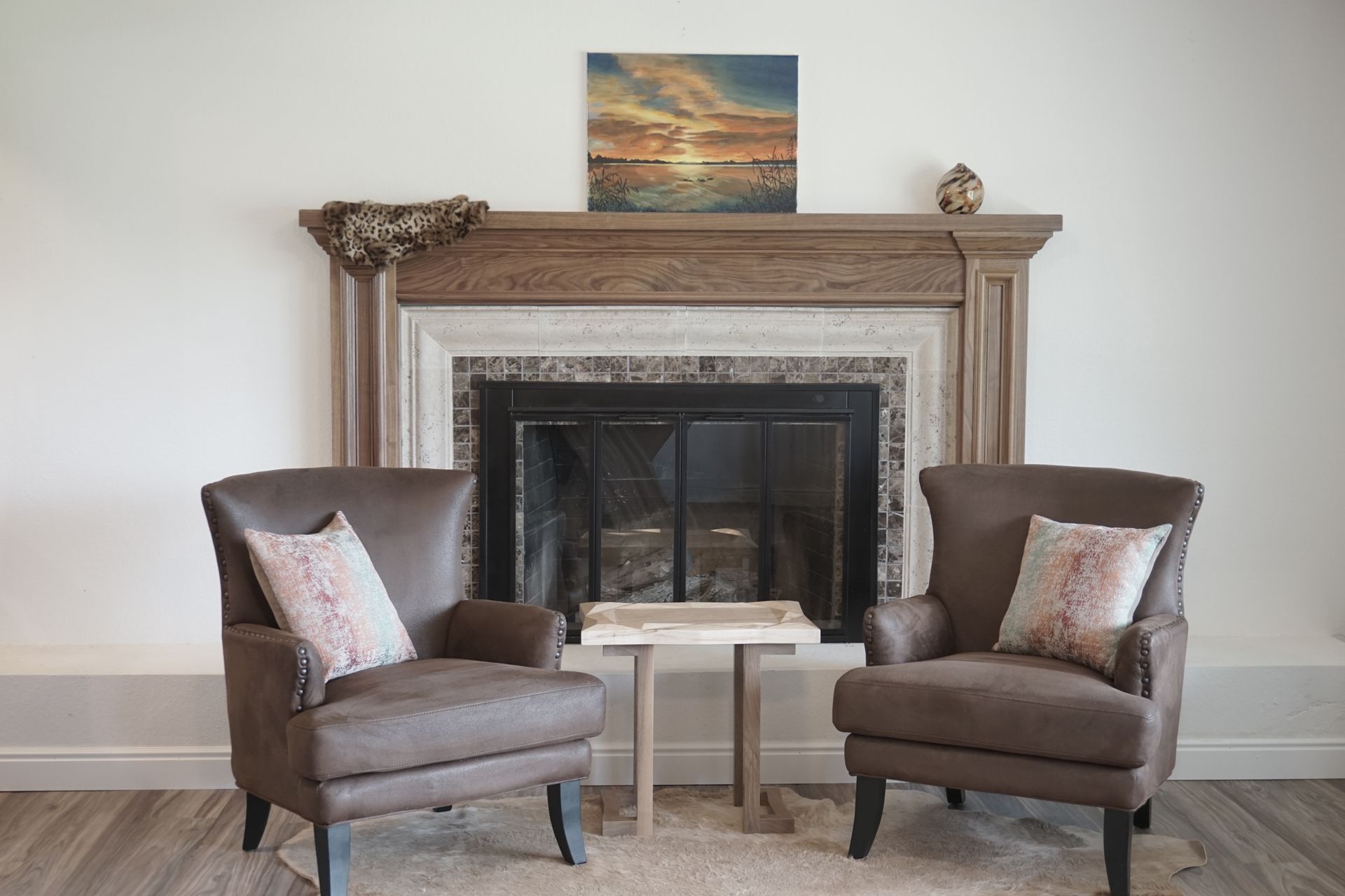 Two brown armchairs face a fireplace with a painting above. A small table sits between the chairs.