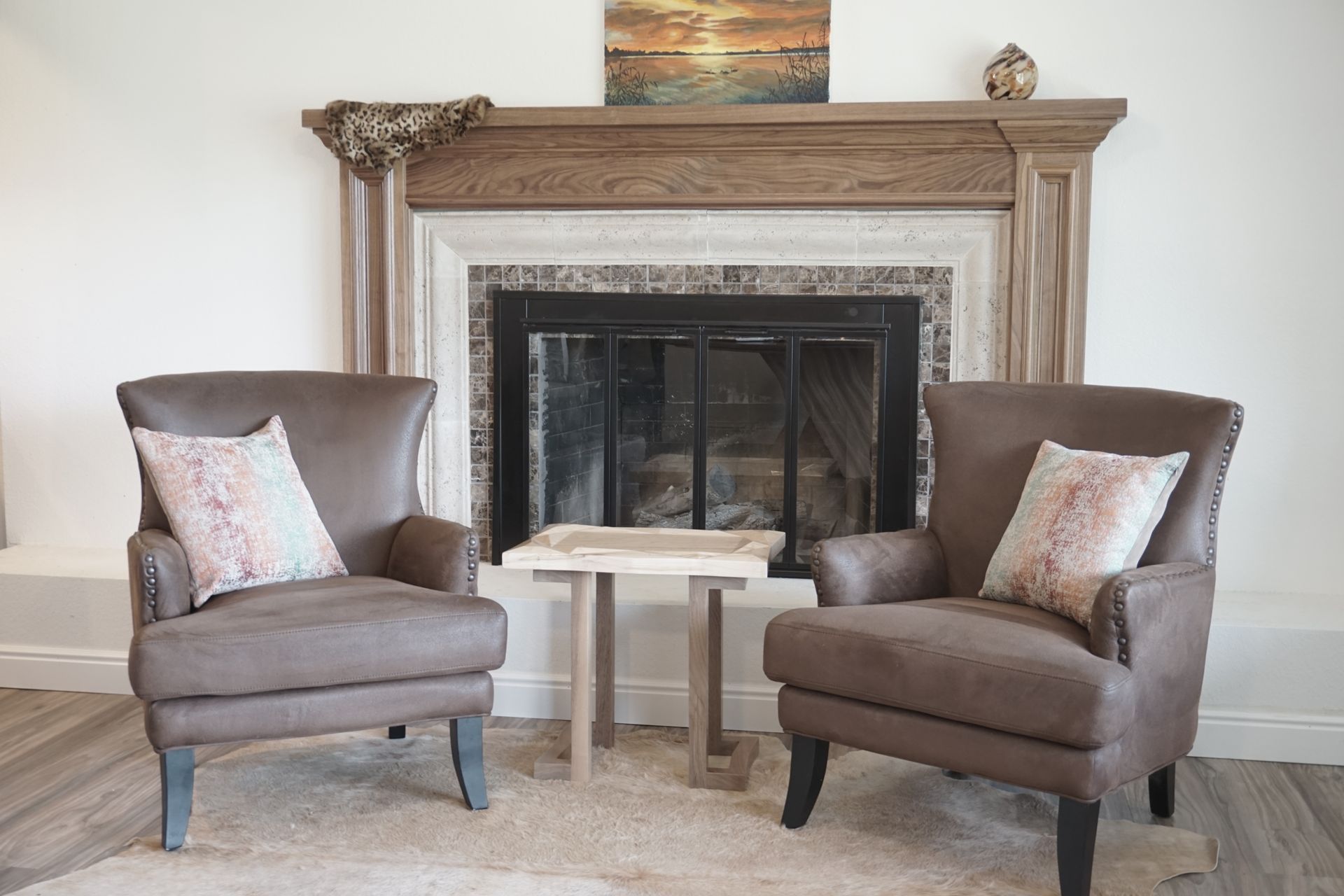 Two brown armchairs face a fireplace, with a small table between them. Beige and brown tones.