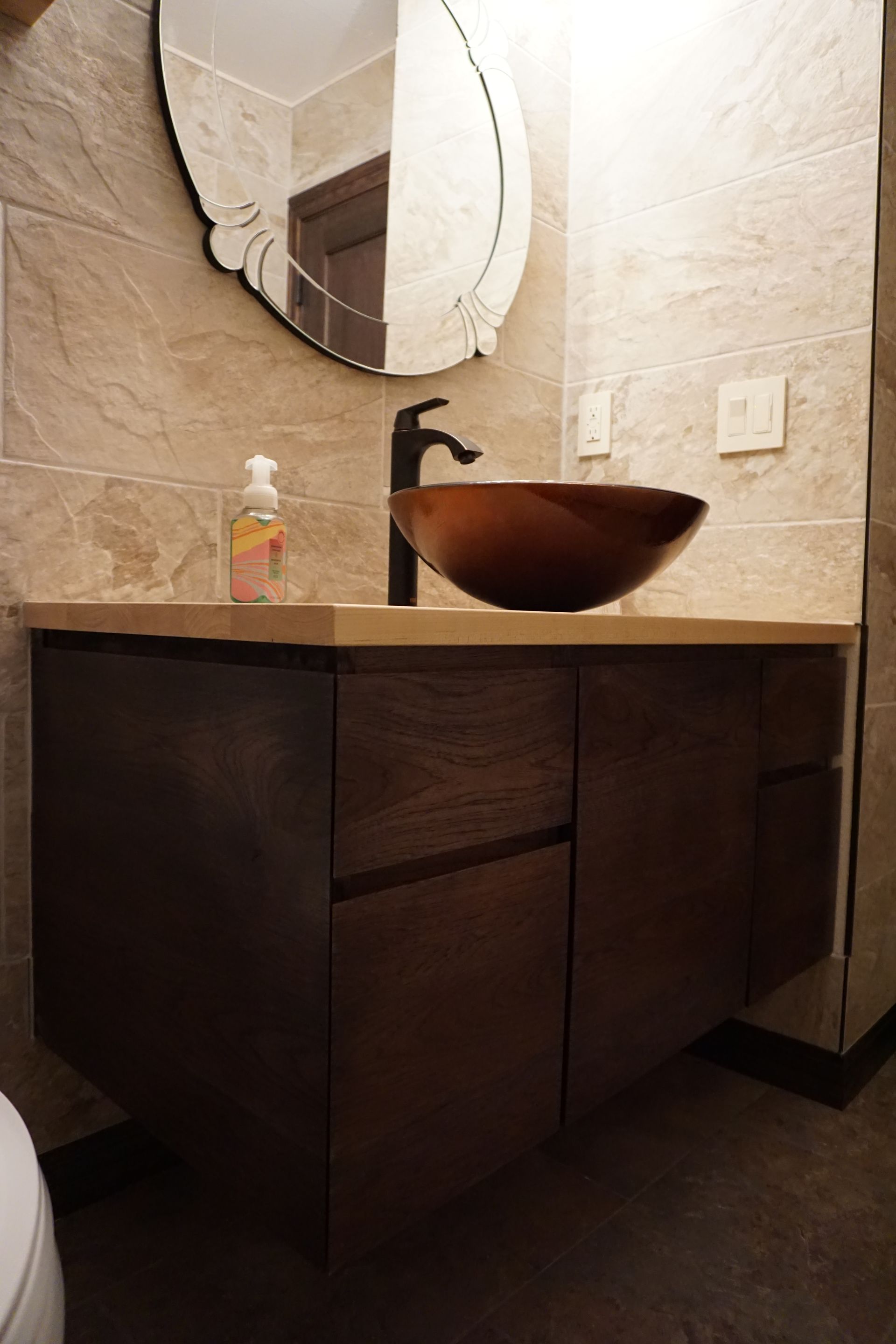 Wooden vanity with bowl sink, black faucet, and oval mirror in a bathroom with beige tile walls.